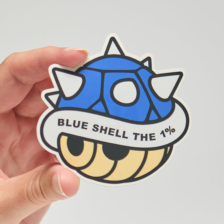 Sticker with blue shell from Mario Brothers that reads, "Blue shell the 1%" held by a hand on a neutral background