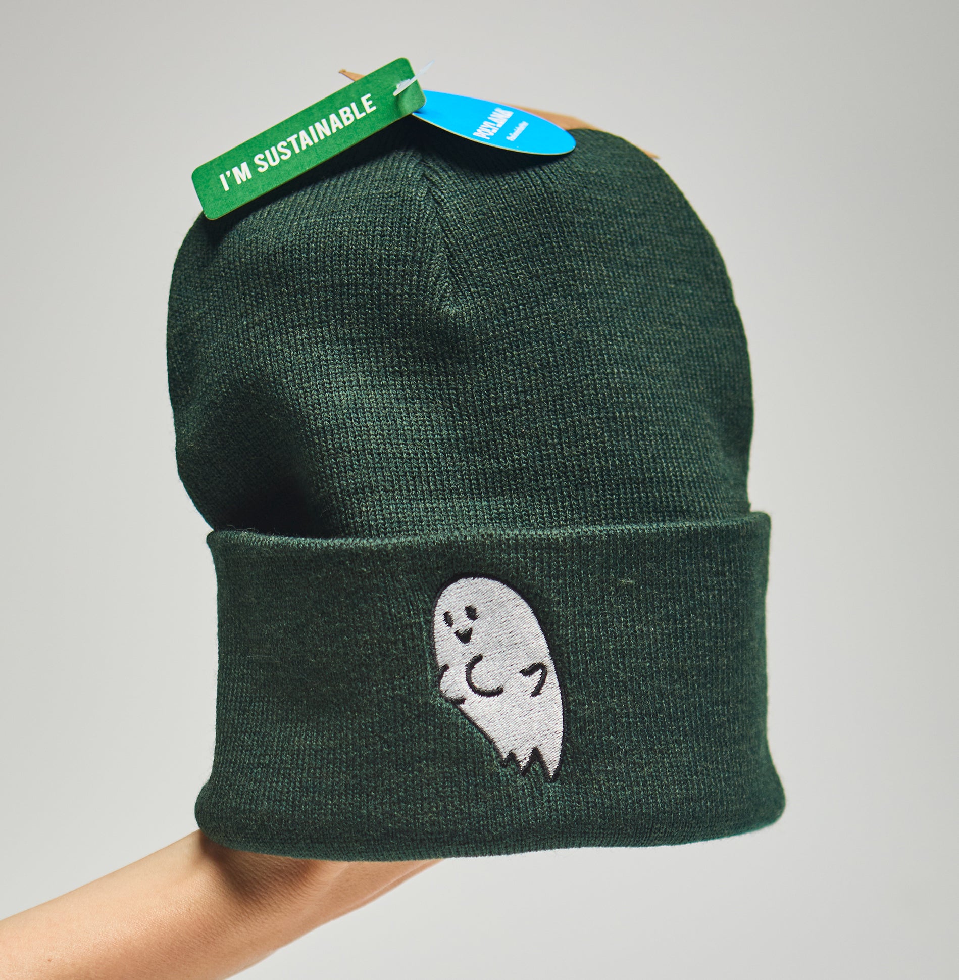 Green beanie with a ghost with boobs design and 'I'm Sustainable' tag on a neutral background