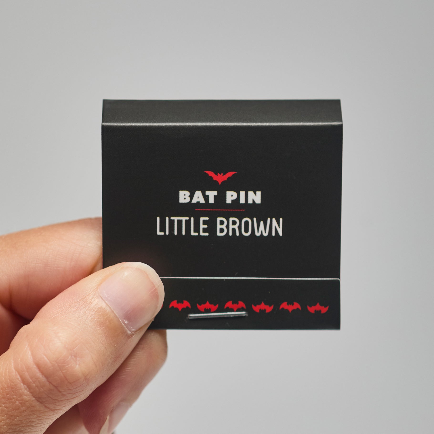 A black match book with 'Bat Pin Little Brown' text held by a hand against a neutral background