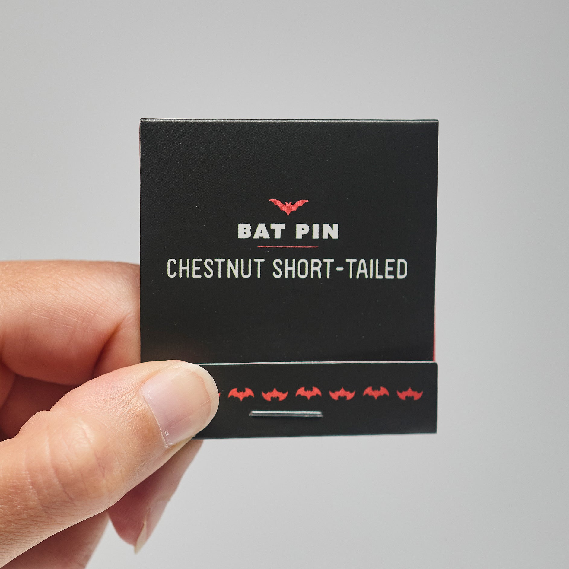 A black match book with 'Bat Pin Chestnut short-tailed' text held by a hand against a neutral background