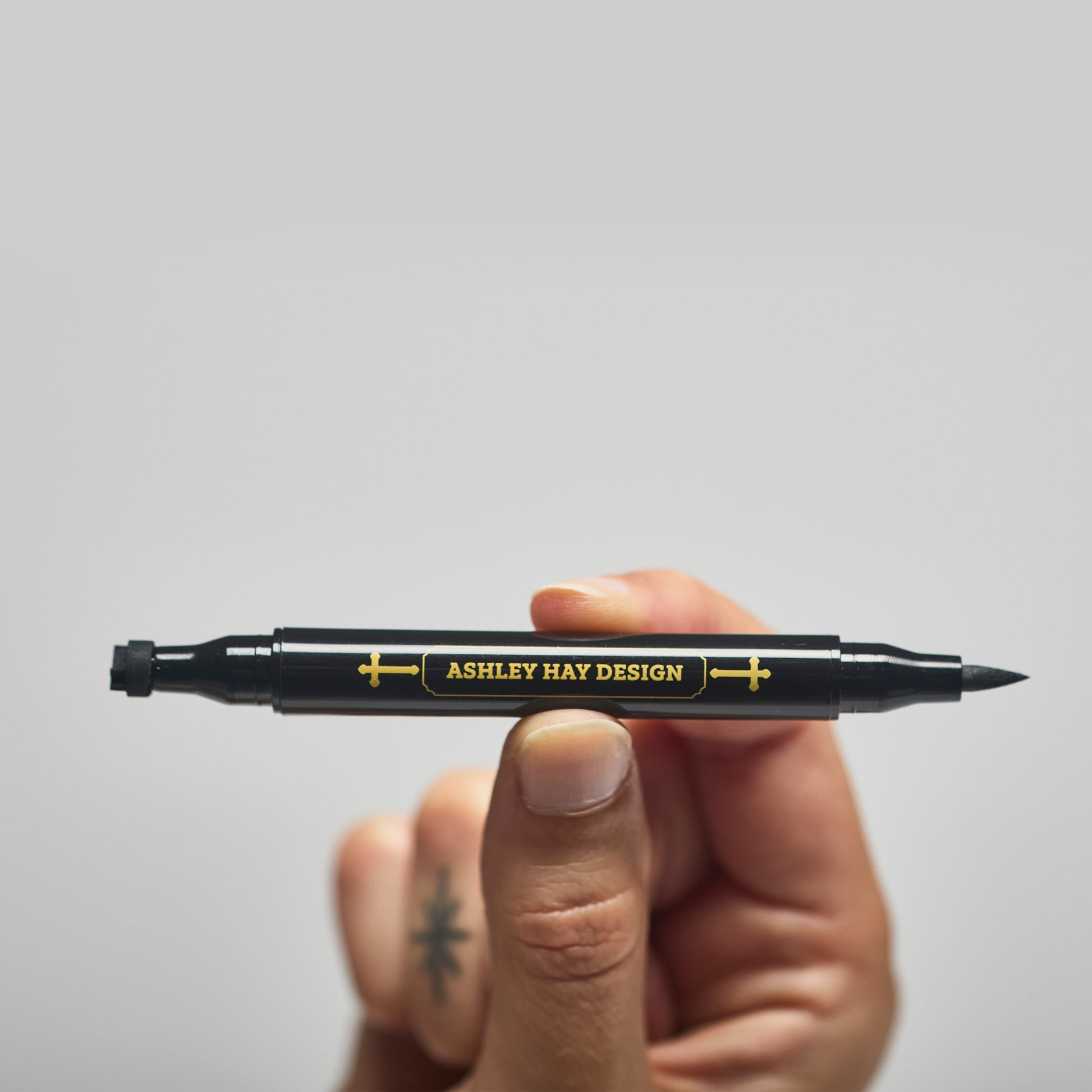 Hand holding a black makeup tube with a brush on one side and a stamp on the other side against a gray background.