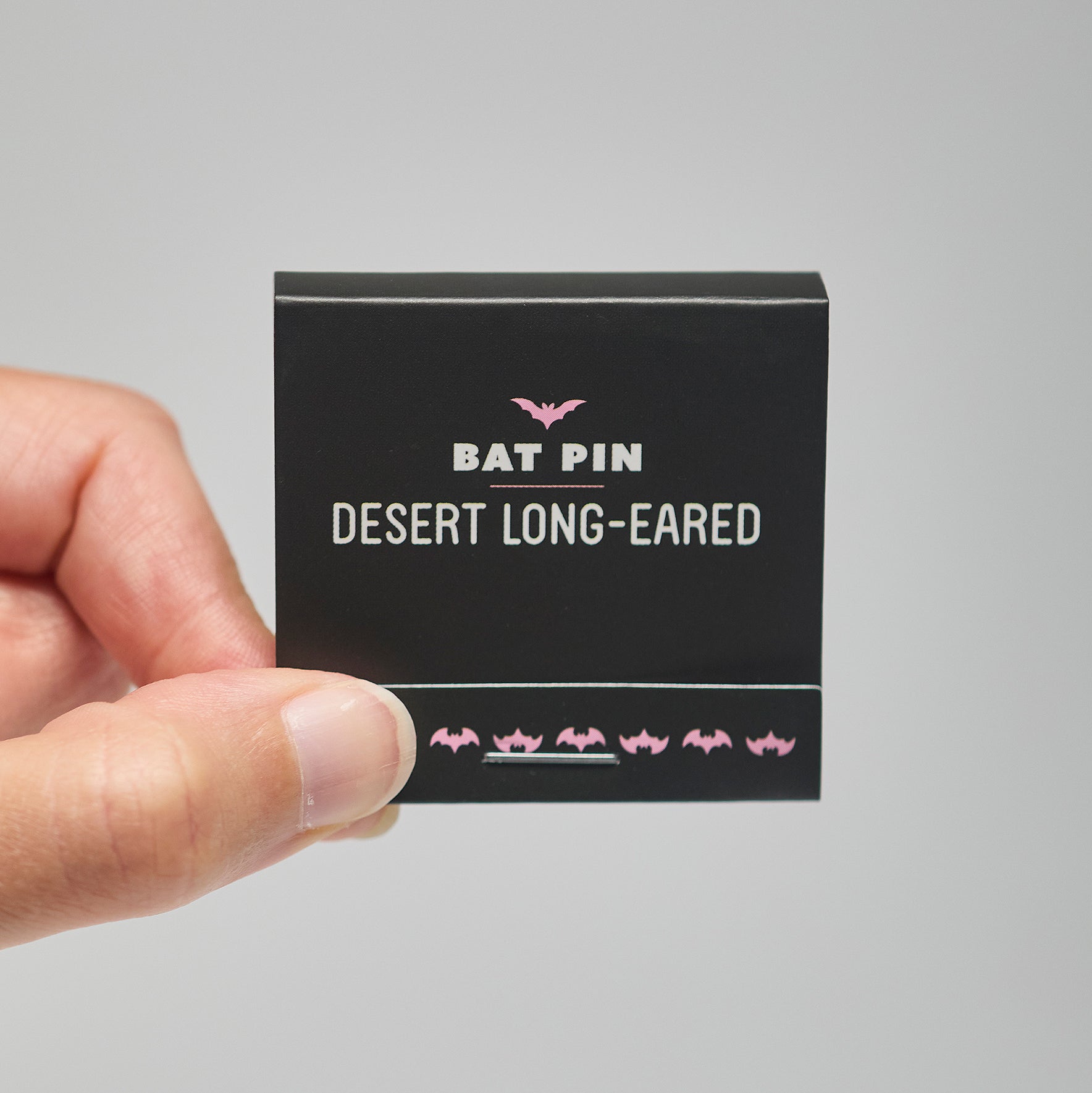 A black match book with 'Bat Pin Desert long-eared' text held by a hand against a neutral background