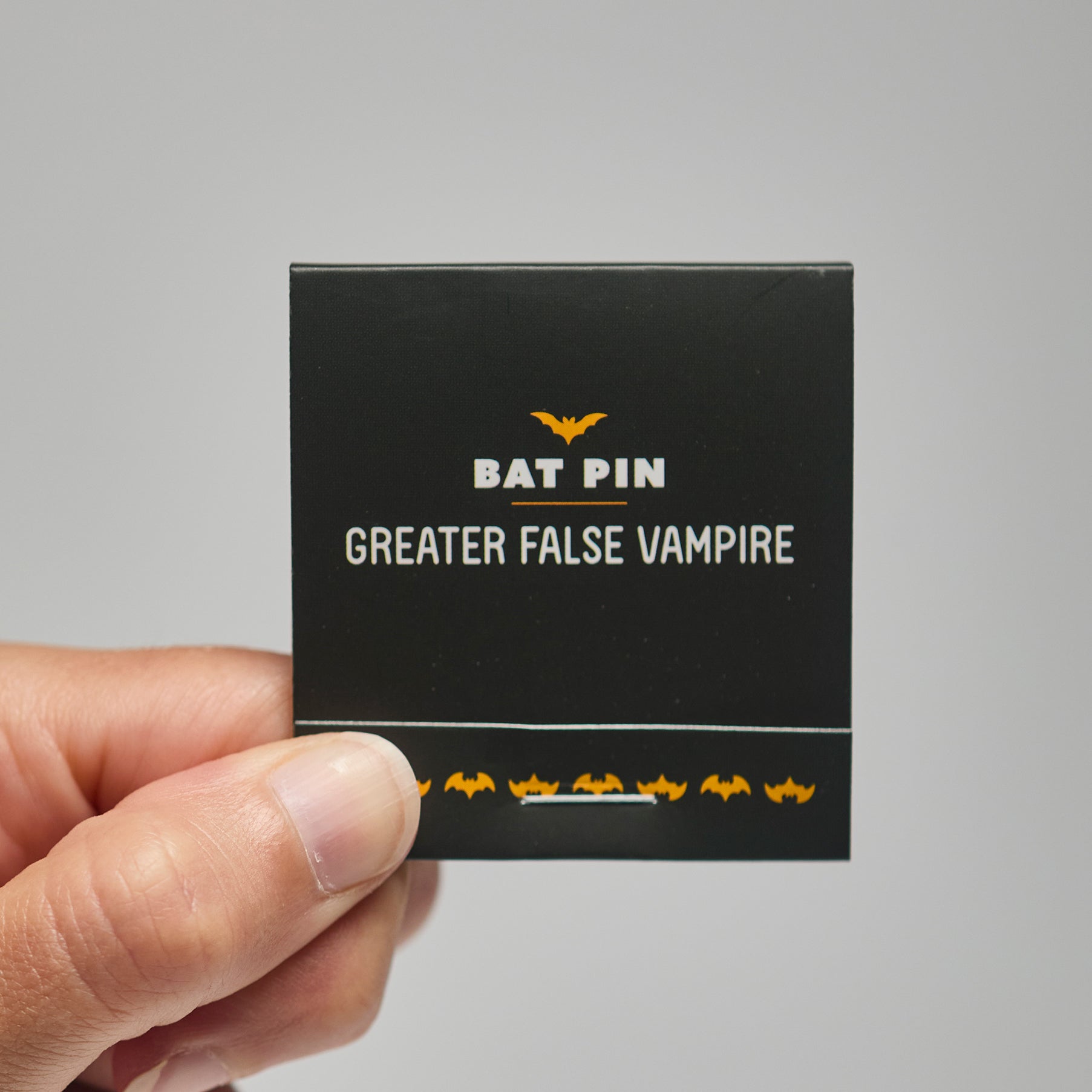 A black match book with 'Bat Pin Greater False Vampire' text held by a hand against a neutral background