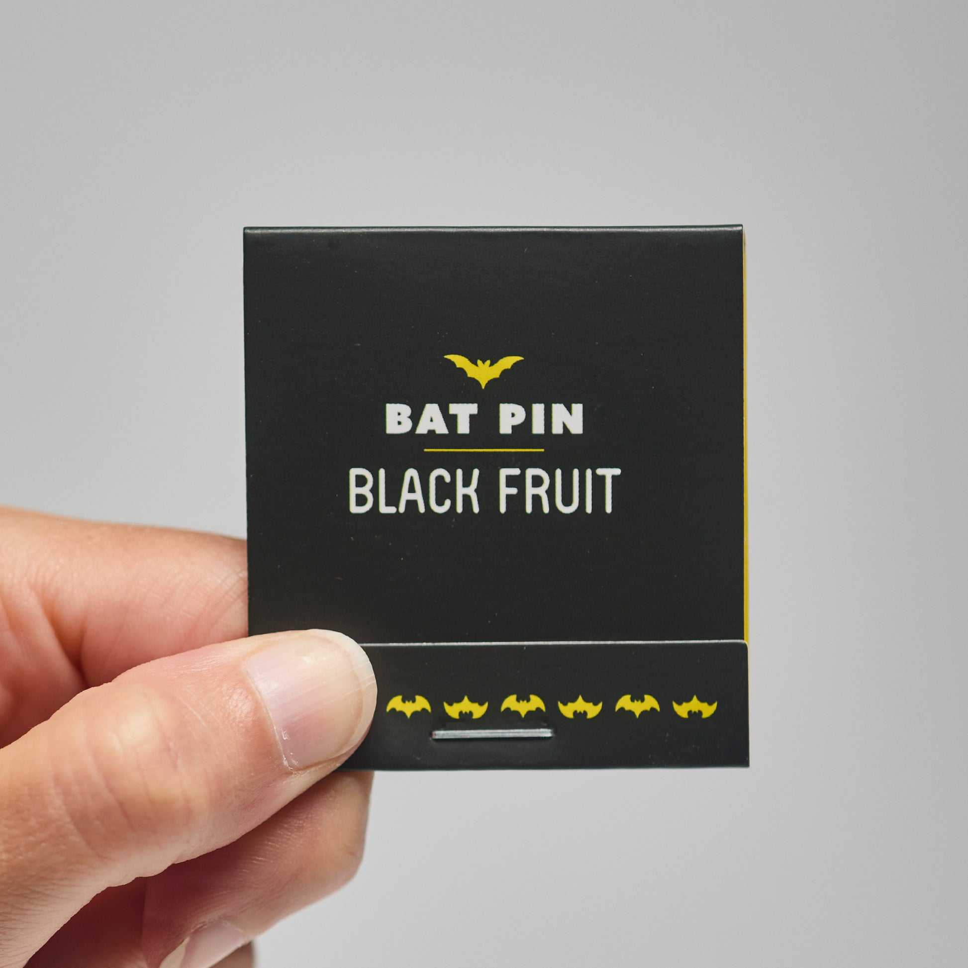 A black match book with 'Bat Pin Black Fruit' text held by a hand against a neutral background