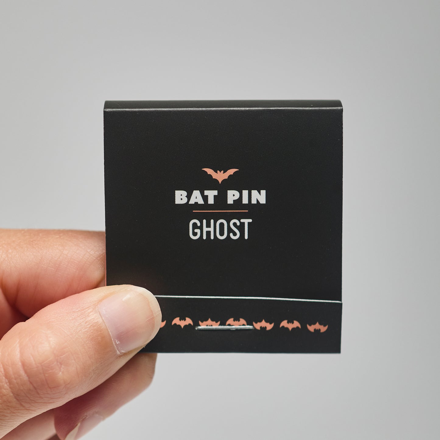 A black match book with 'Bat Pin Ghost' text held by a hand against a neutral background