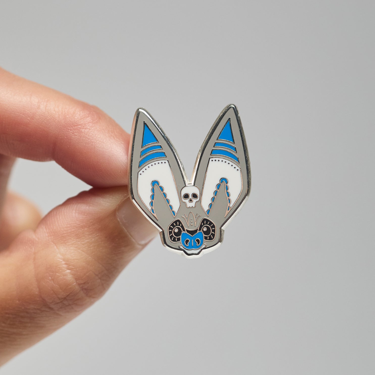 Enamel pin of a grey long-eared bat held by a hand against a neutral background