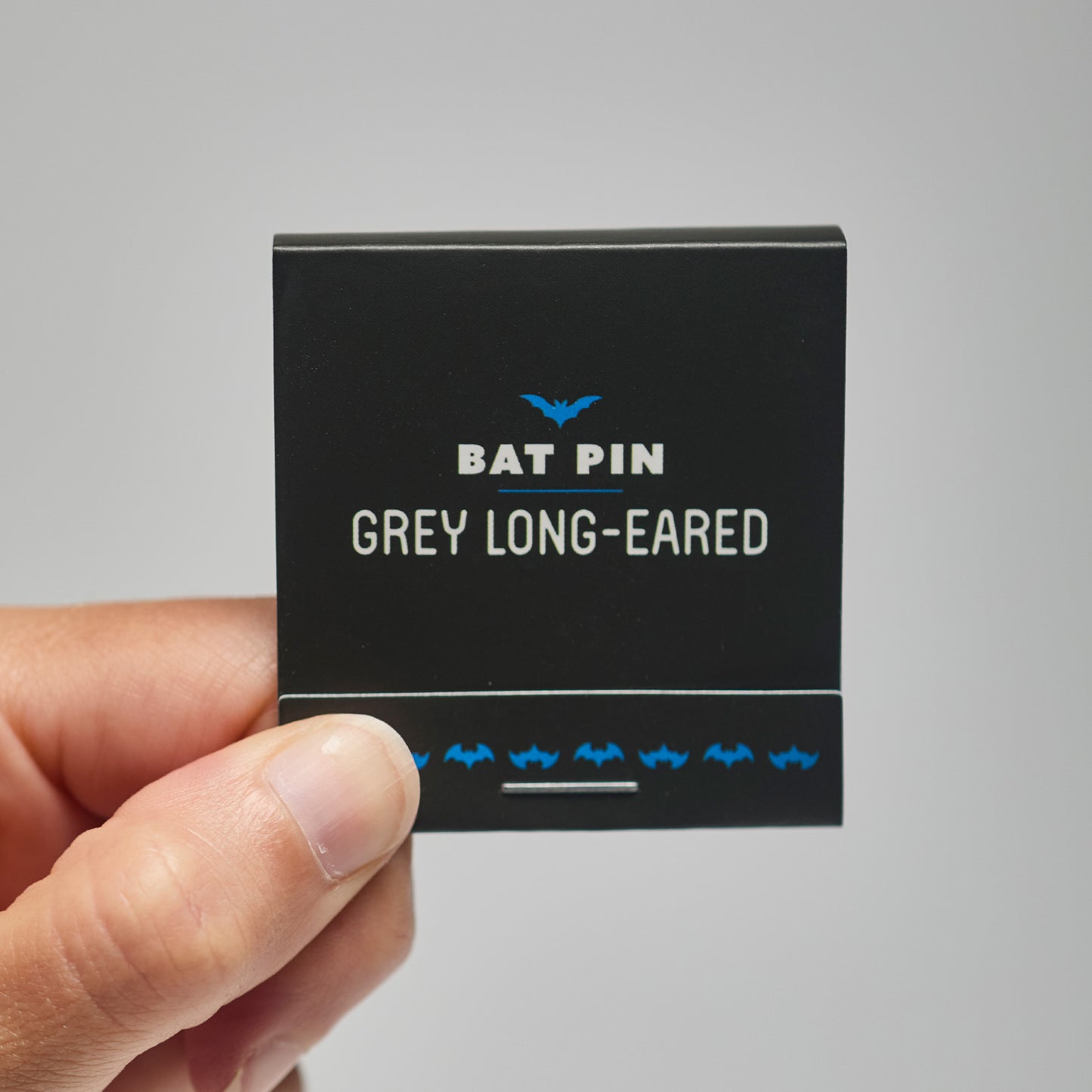 A black match book with 'Bat Pin grey long-eared' text held by a hand against a neutral background