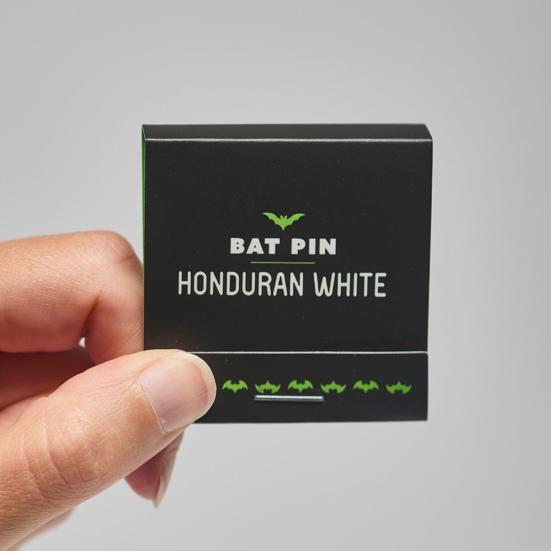 A black match book with 'Bat Pin Honduran white' text held by a hand against a neutral background