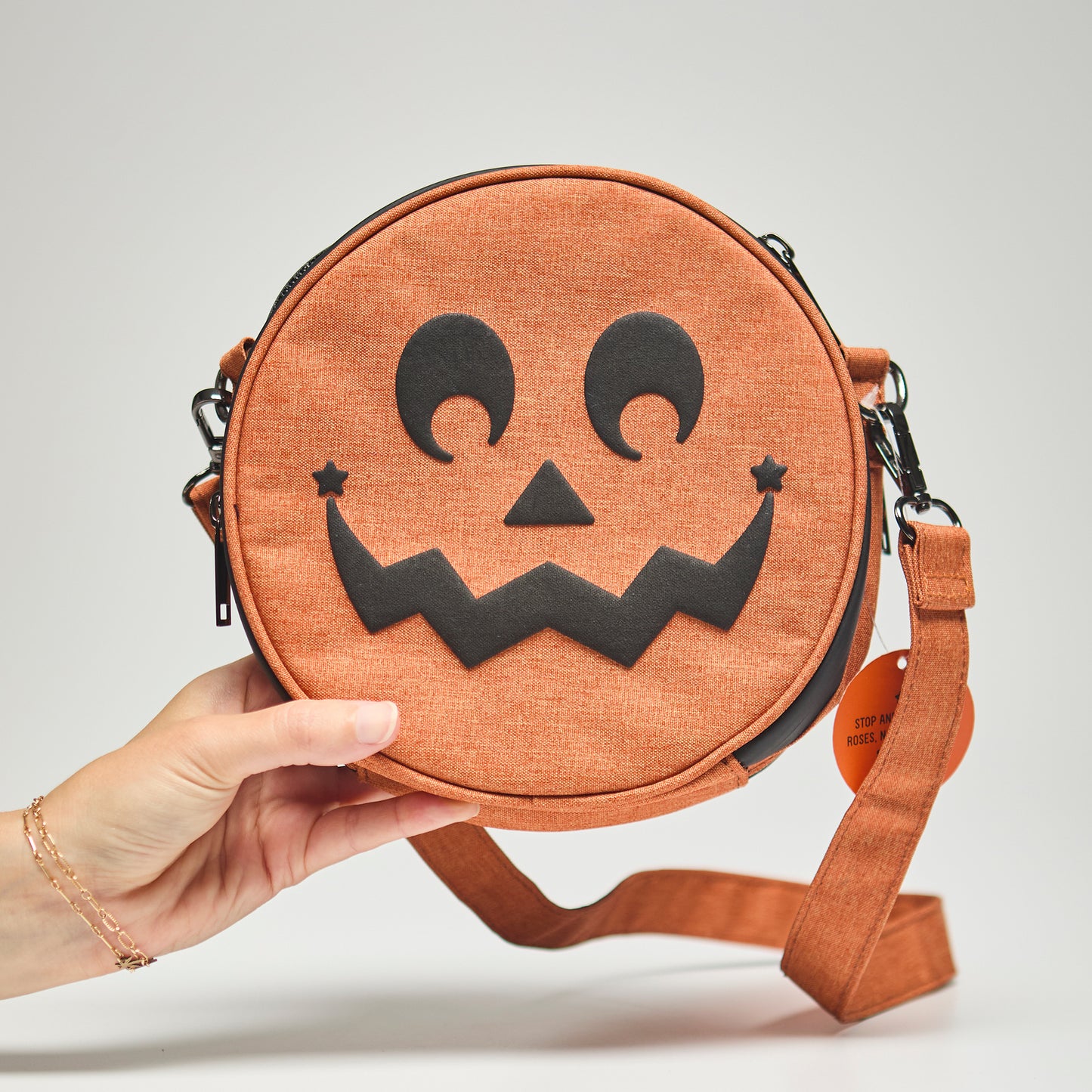 Round orange bag with a jack-o'-lantern face design held by a hand on a neutral background