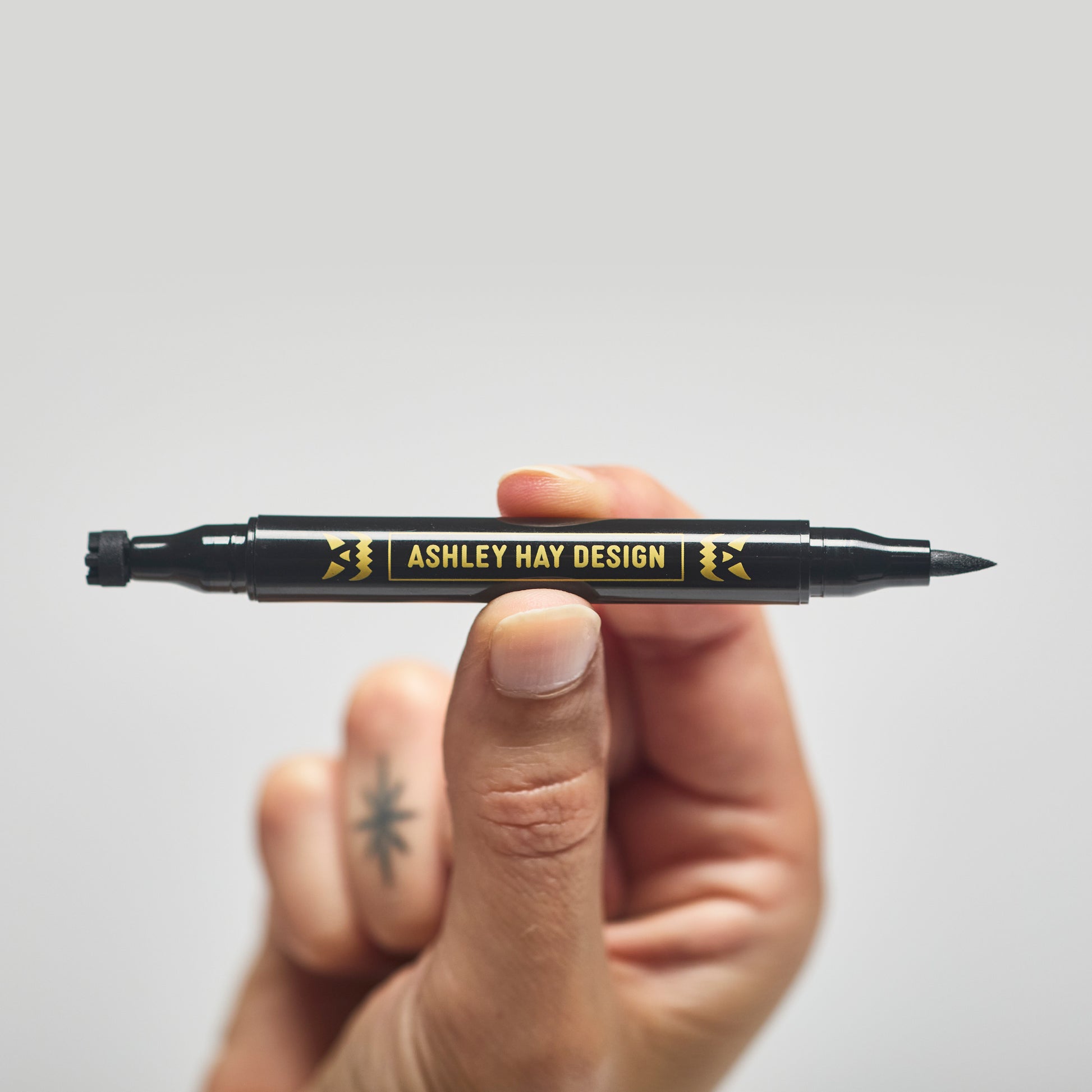 Hand holding a black makeup tube with a brush on one side and a stamp on the other side against a gray background.