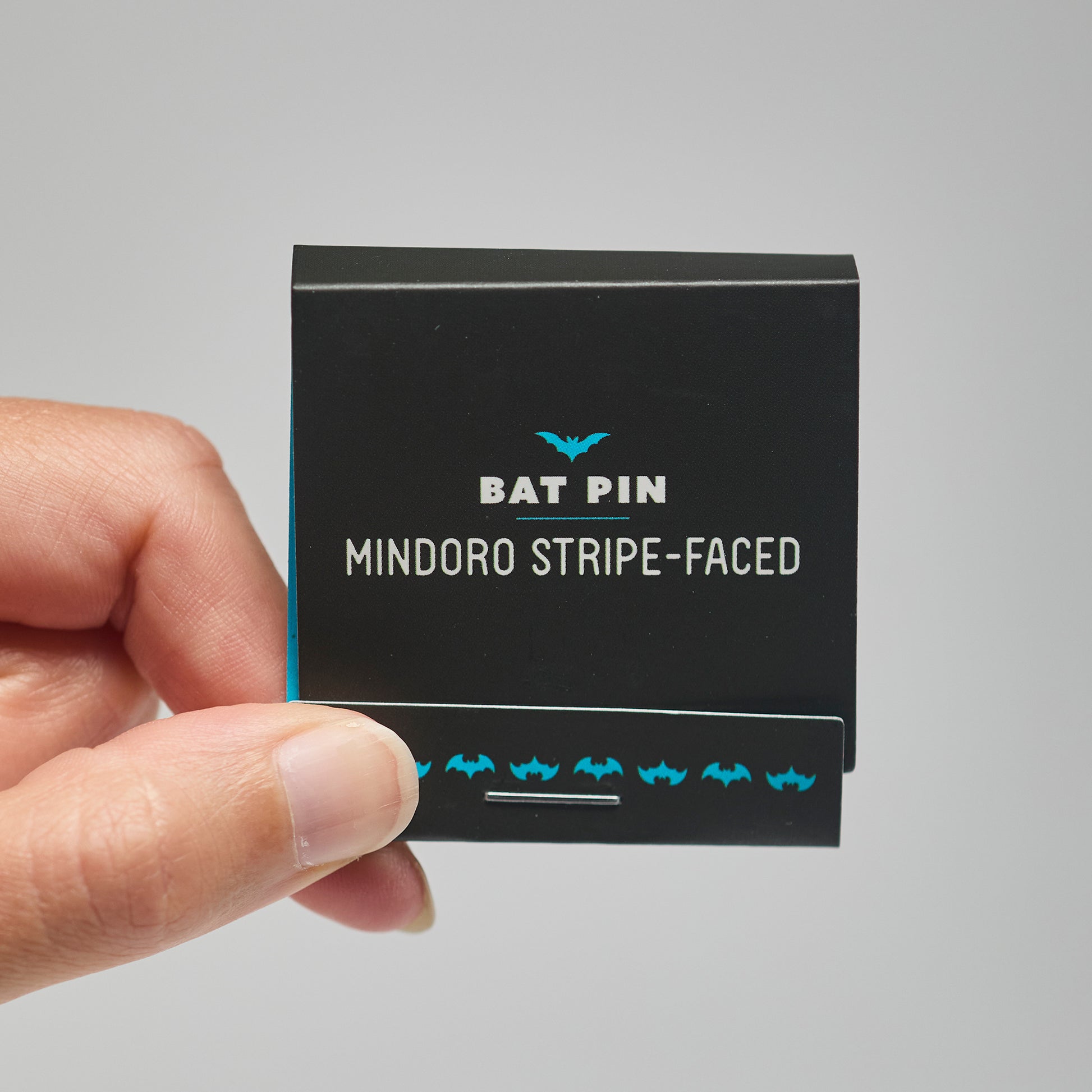 A black match book with 'Bat Pin mindoro stripe-faced' text held by a hand against a neutral background