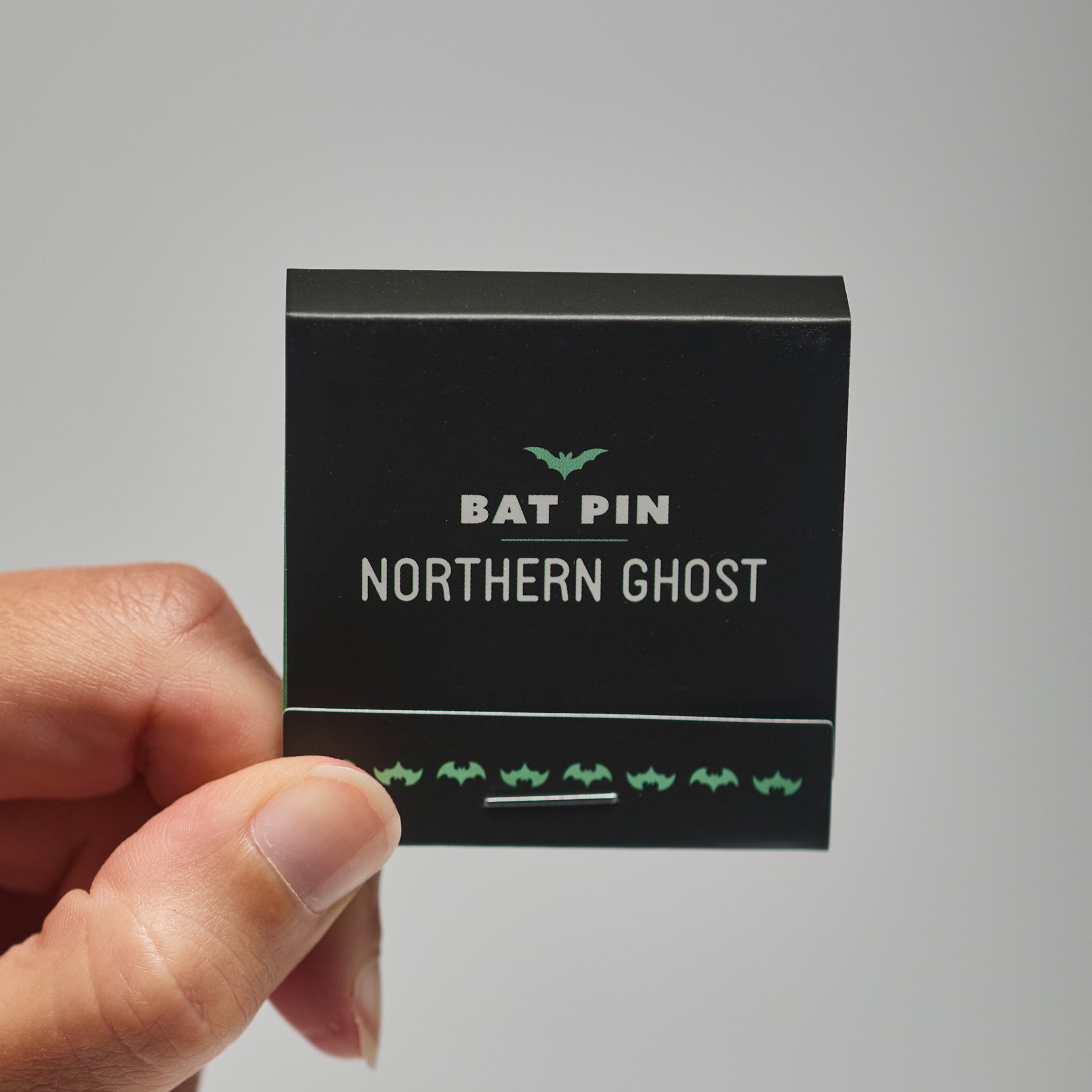 A black match book with 'Bat Pin Northern ghost' text held by a hand against a neutral background