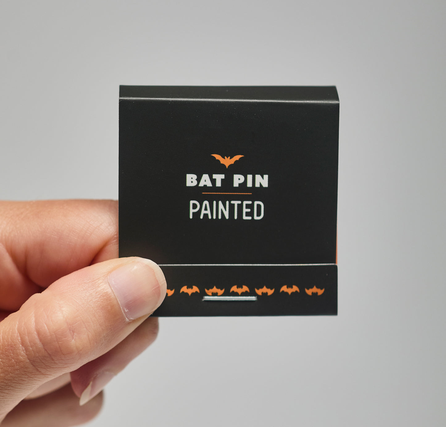 A black match book with 'Bat Pin Painted' text held by a hand against a neutral background
