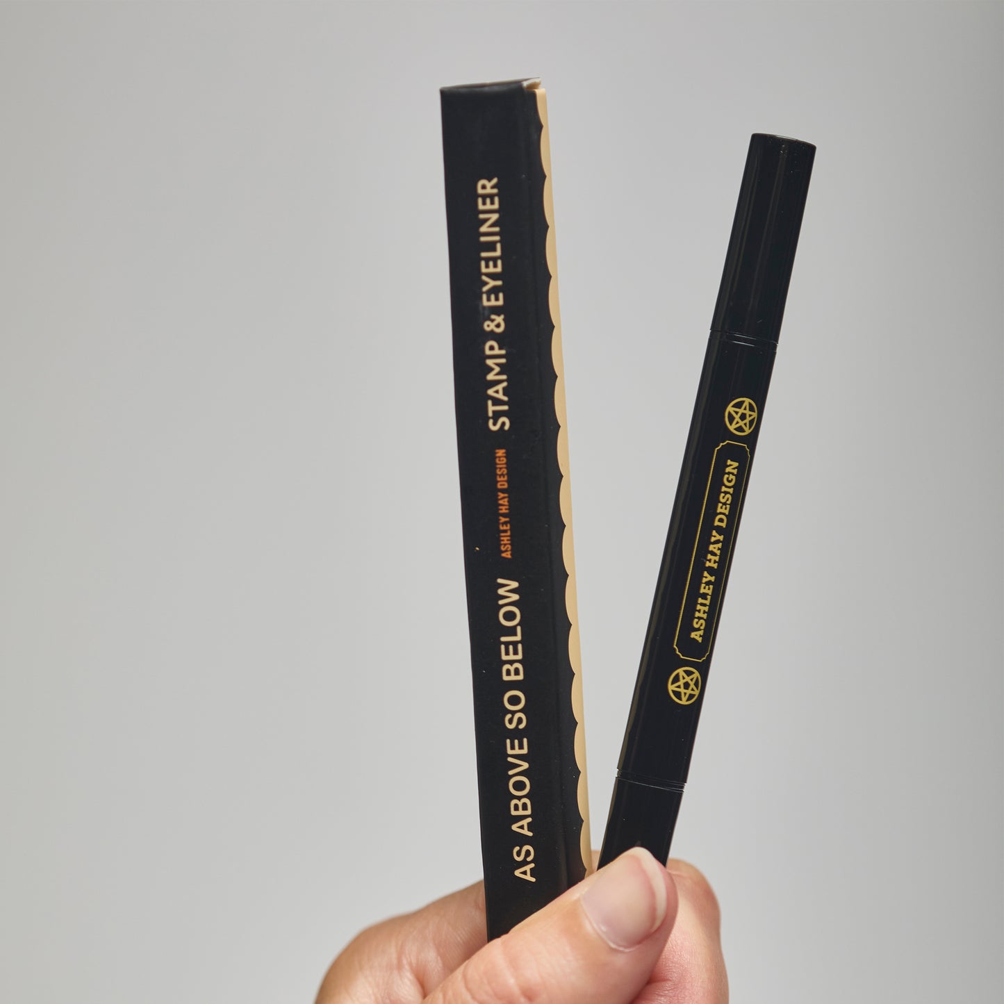 Hand holding a black stamp and eyeliner packaging with visible branding against a gray background