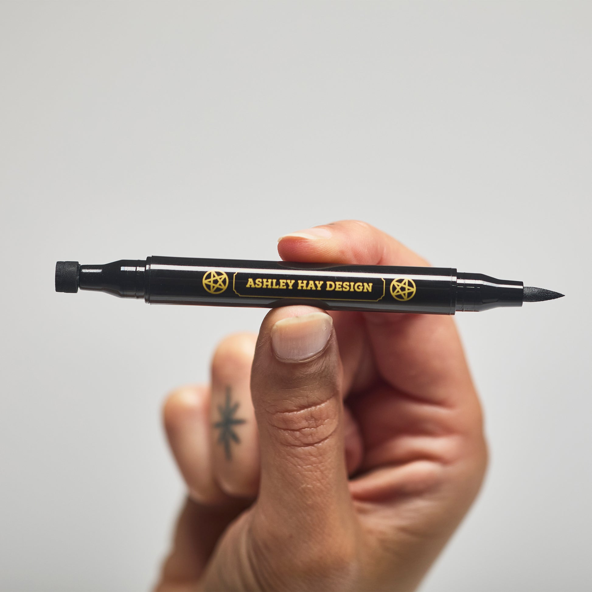 Hand holding a black makeup tube with a brush on one side and a stamp on the other side against a gray background.