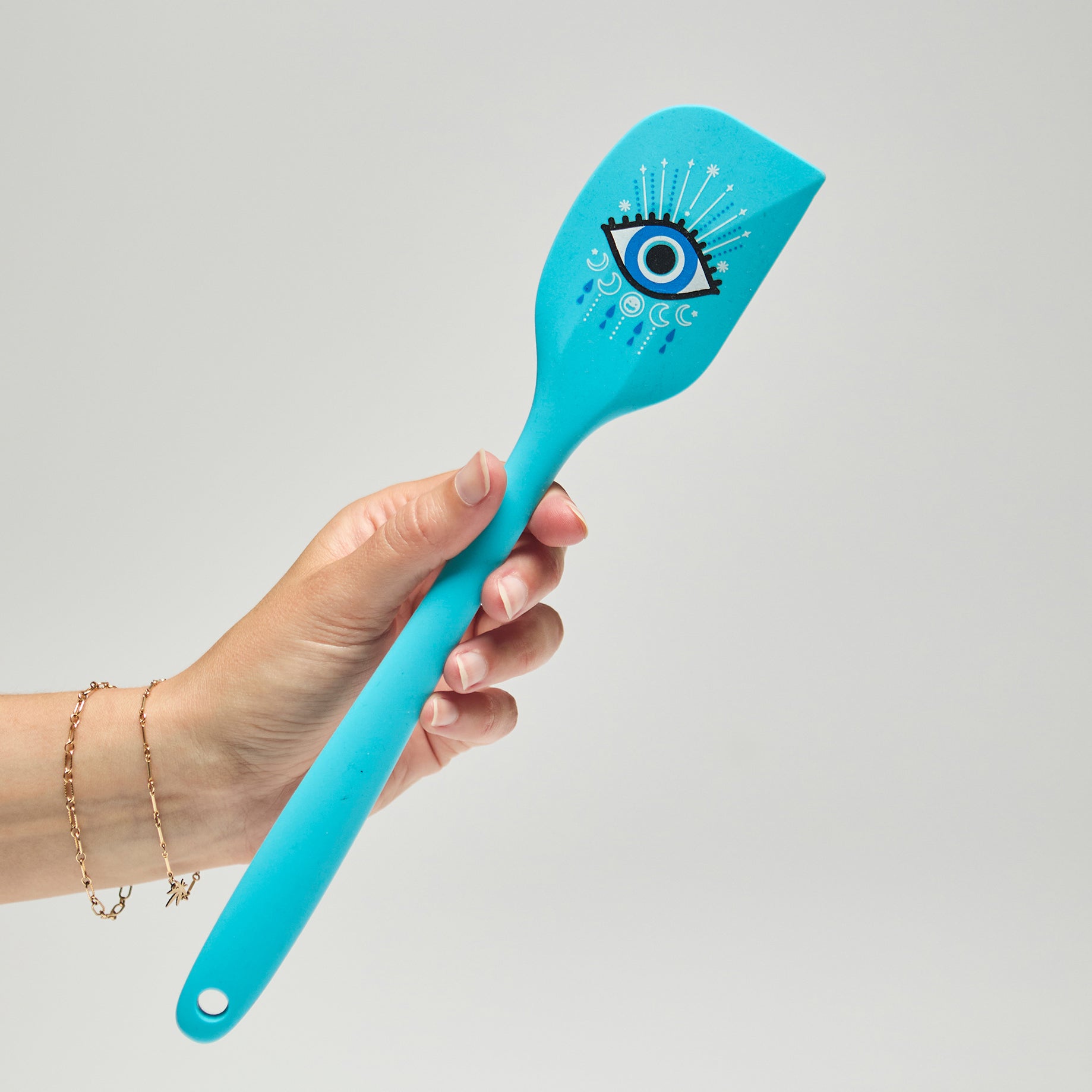 A hand holding a bright blue spatula with an evil eye illustration against a neutral bckground