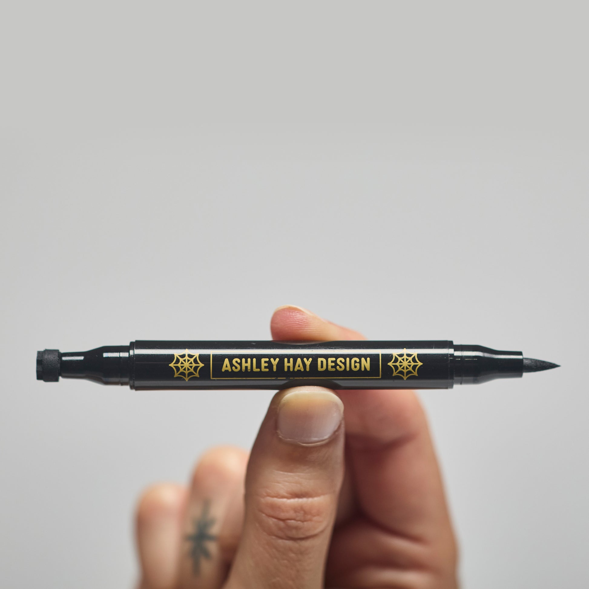 Hand holding a black makeup tube with a brush on one side and a stamp on the other side against a gray background.