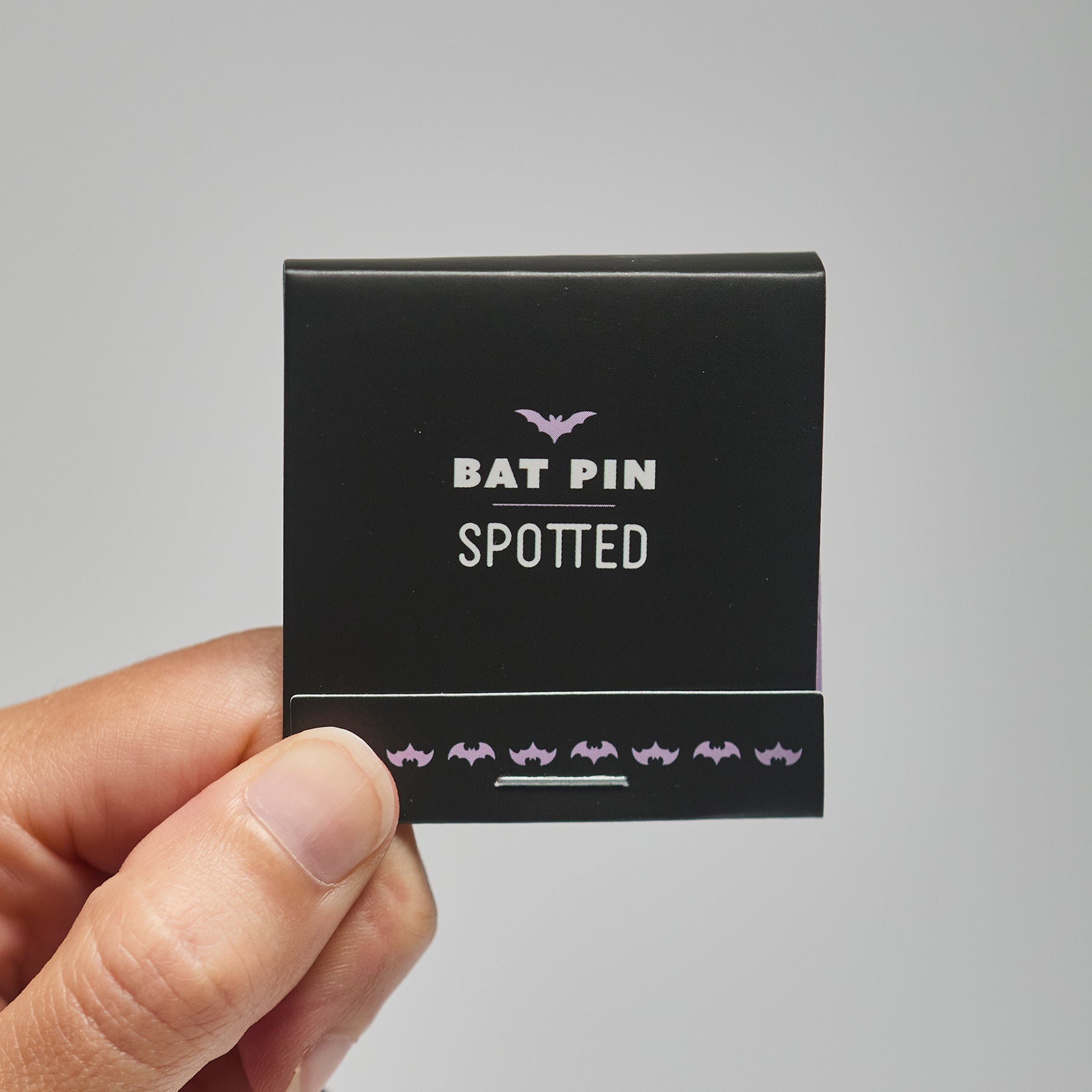 A black match book with 'Bat Pin Spotted' text held by a hand against a neutral background