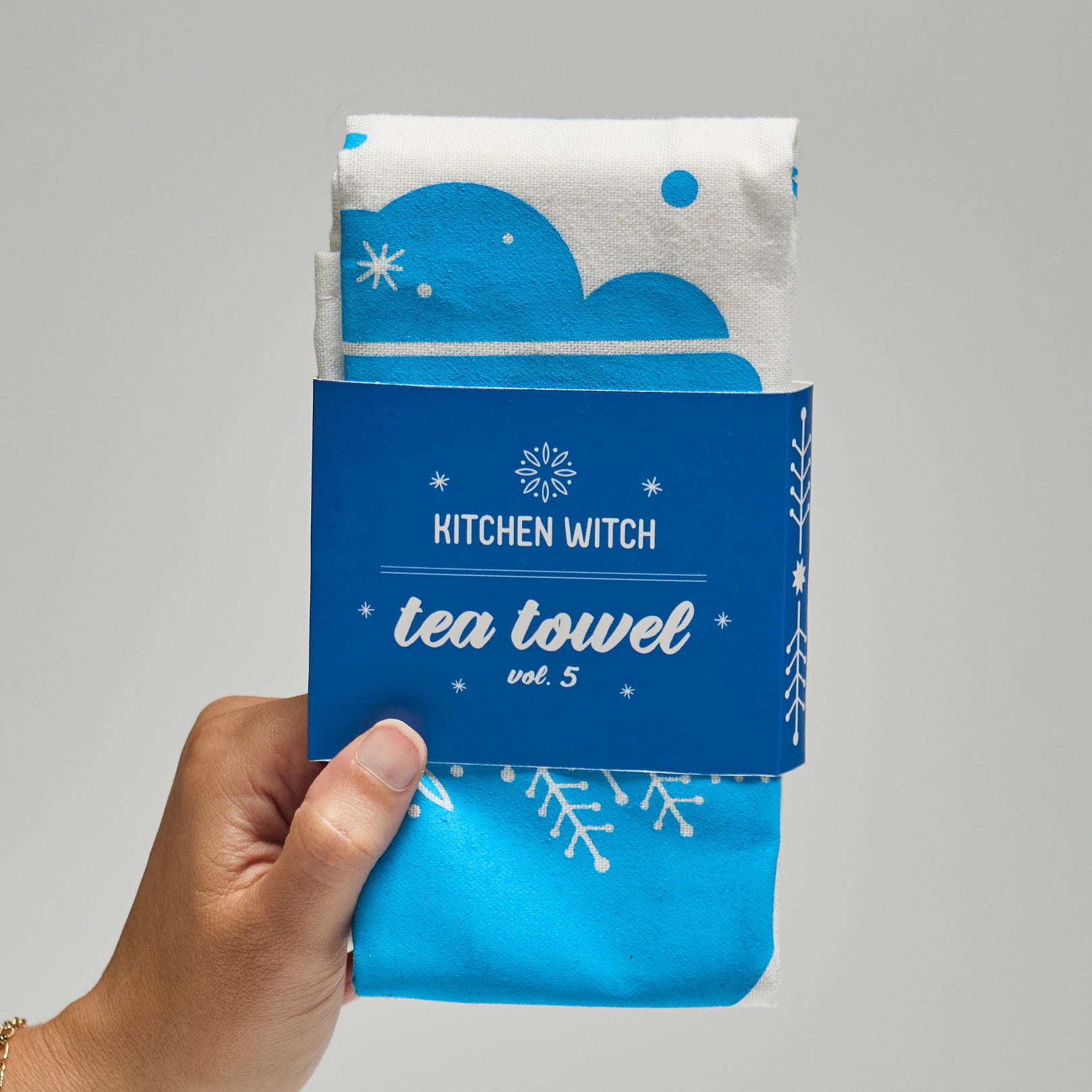 A white and blue tea towel folded with branding reading, 'kitchen witch vol 5' against a neutral background