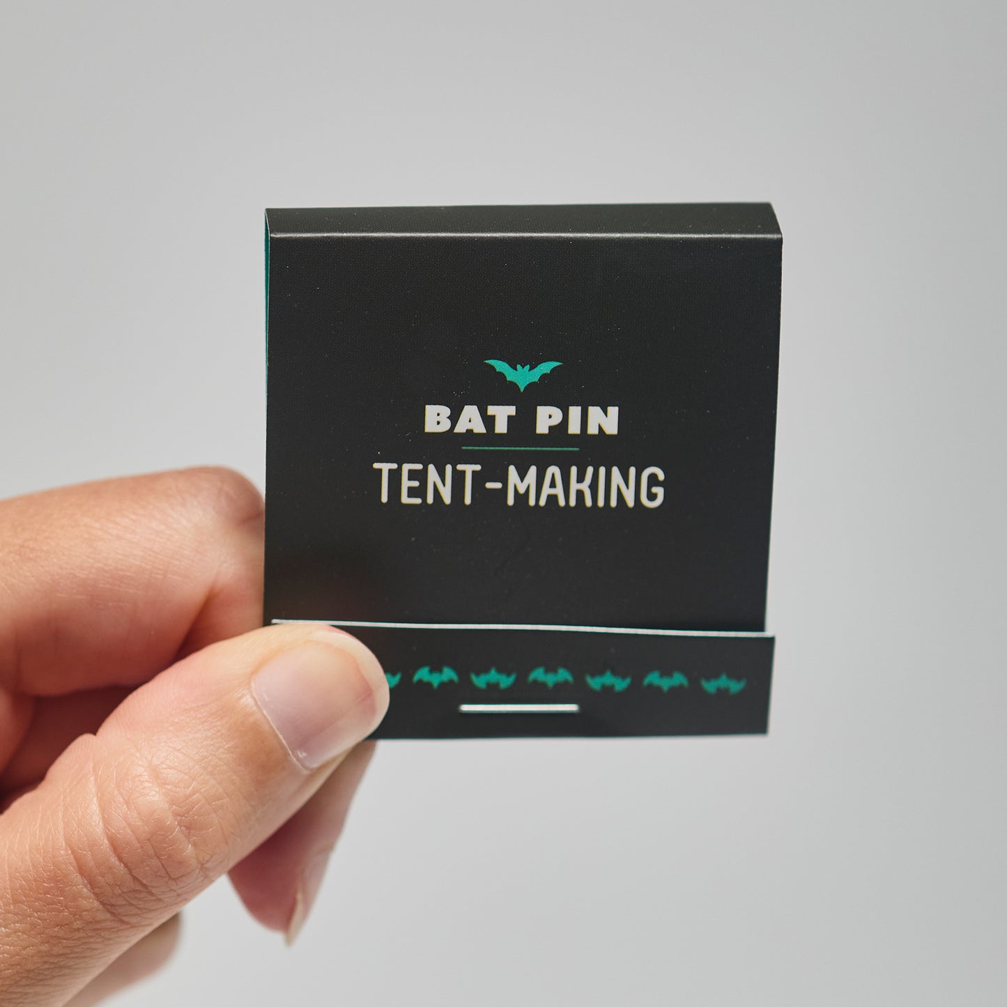 A black match book with 'Bat Pin Tent-making' text held by a hand against a neutral background