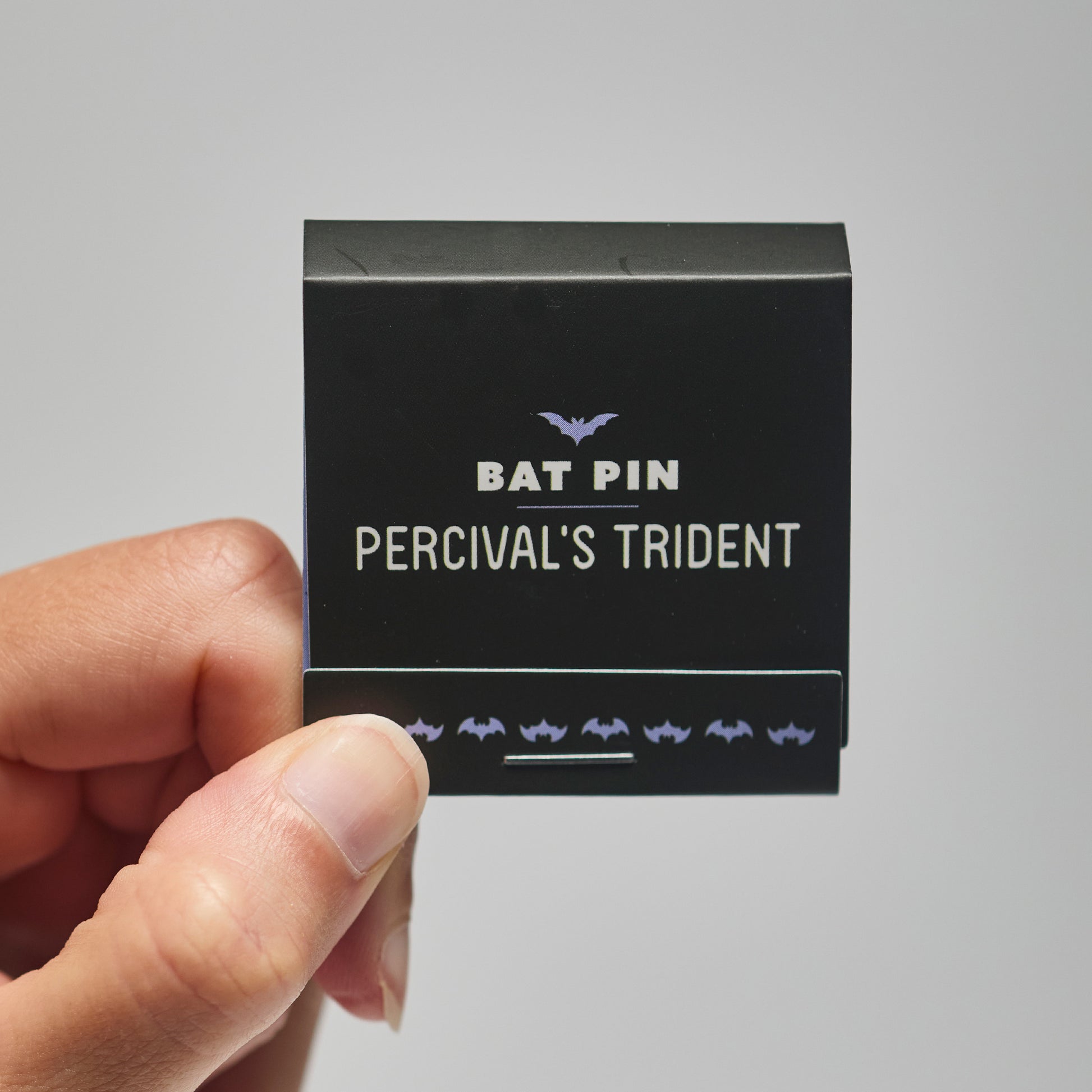 A black match book with 'Bat Pin Pervical's trident' text held by a hand against a neutral background