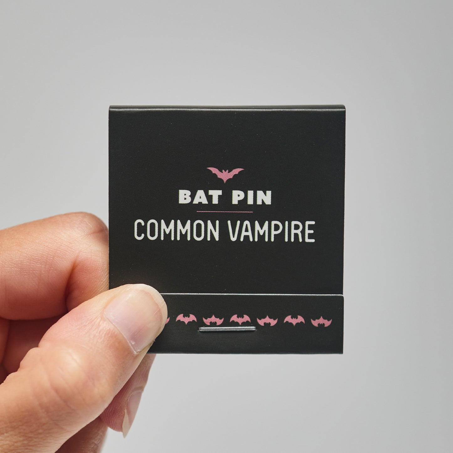 A black match book with 'Bat Pin Common Vampire' text held by a hand against a neutral background