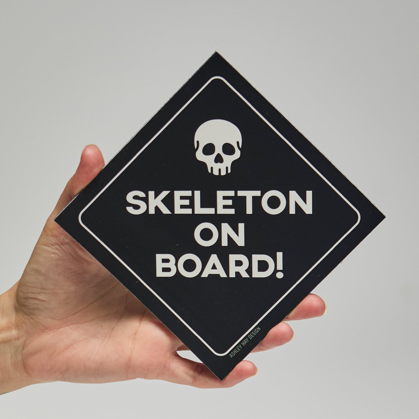 Hand holding a black sign with a skull icon and 'SKELETON ON BOARD!' text on a neutral background