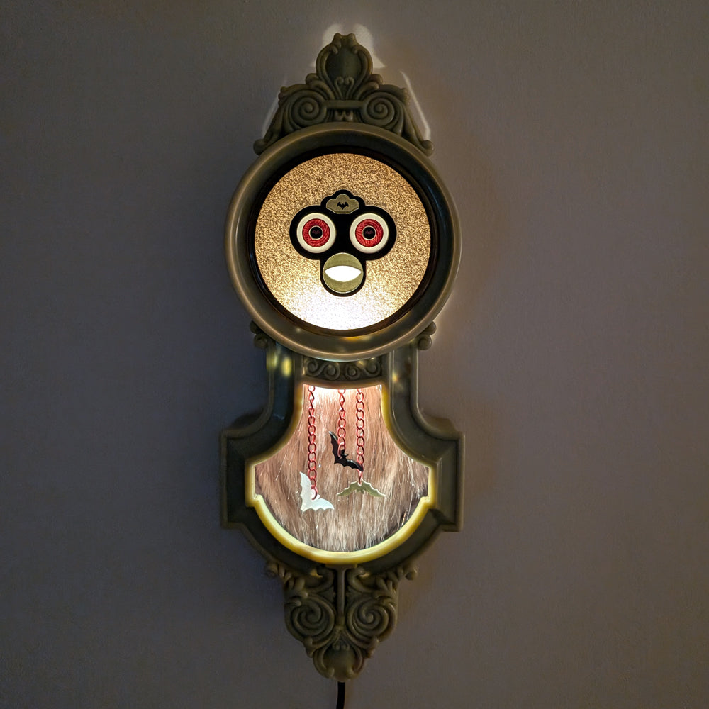 Hanging vintage clock frame with a Furby face lit up against a dark background.
