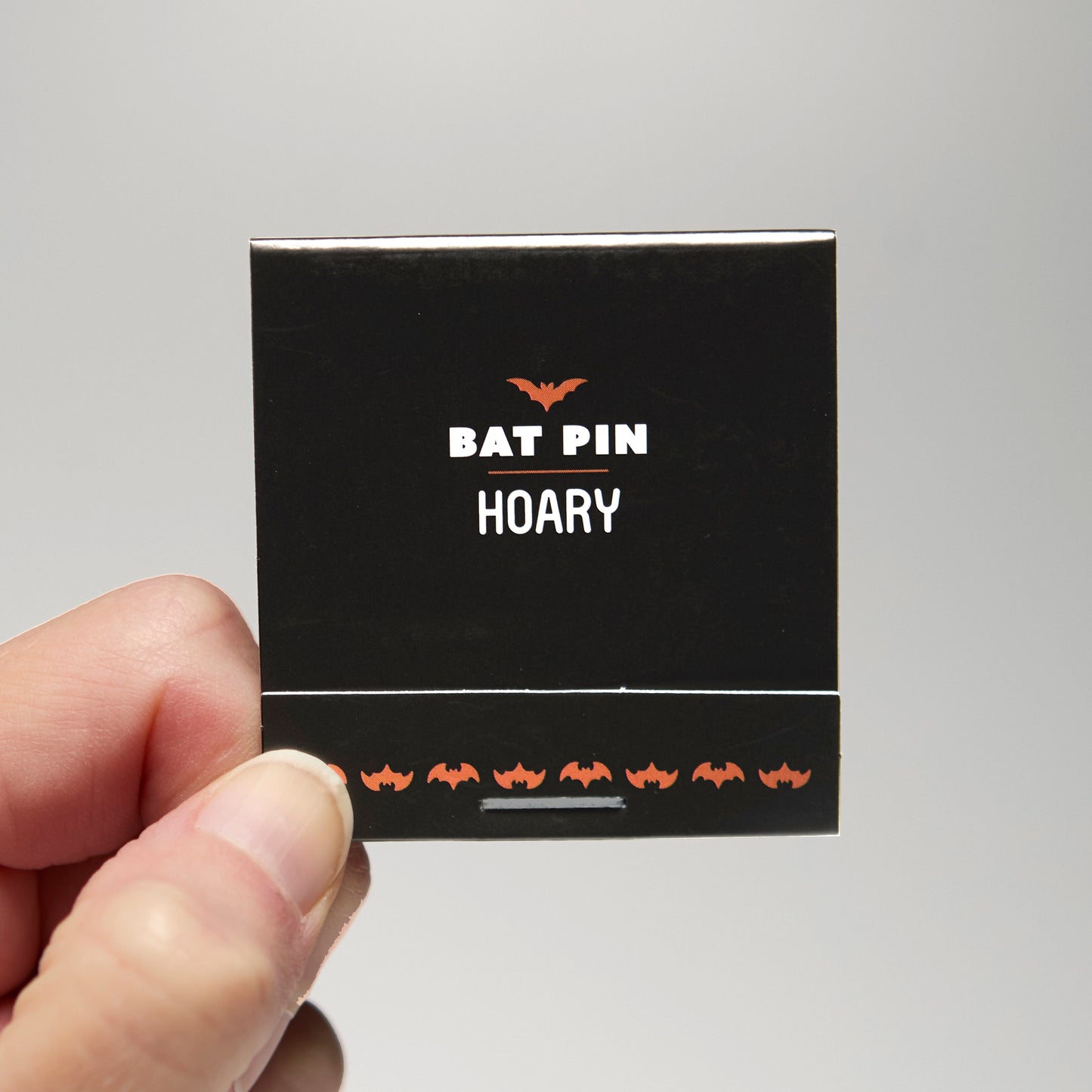 A black match book with 'Bat Pin Hoary' text held by a hand against a neutral background