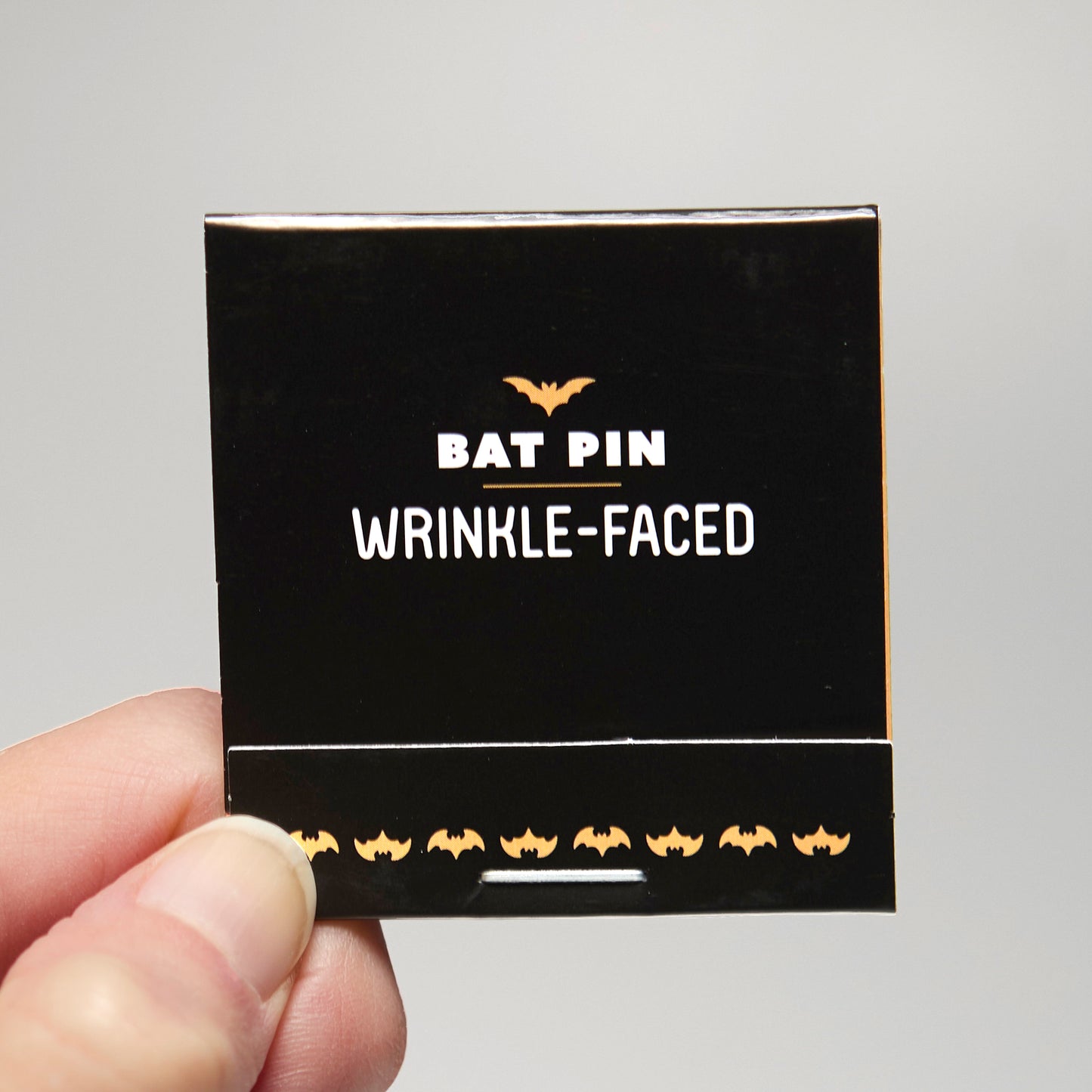 A black match book with 'Bat Pin Wrinkle-faced' text held by a hand against a neutral background