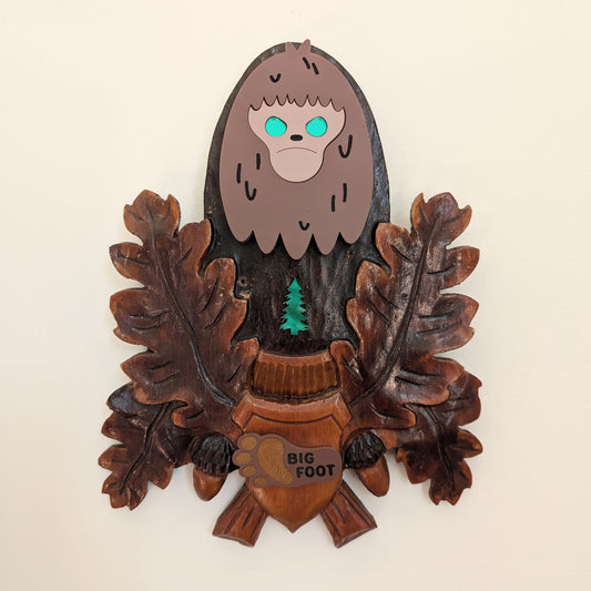 Wooden plaque with a cartoon-like depiction of Bigfoot surrounded by leaves on a beige background