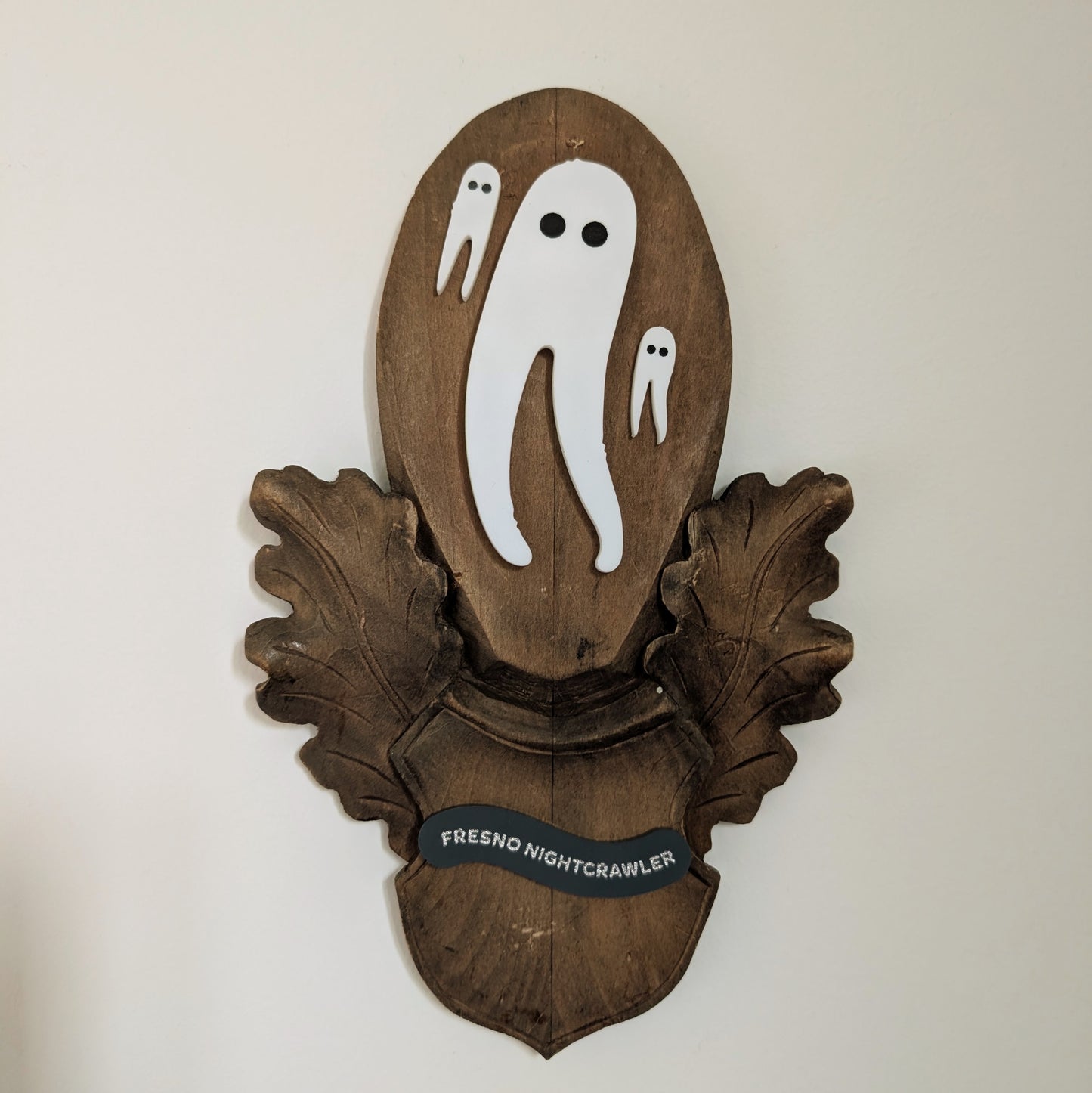 Three acrylic Fresno Nightcrawlers (white leggy cryptids are mounted on a vintage wooden plaque that hangs on a white wall.