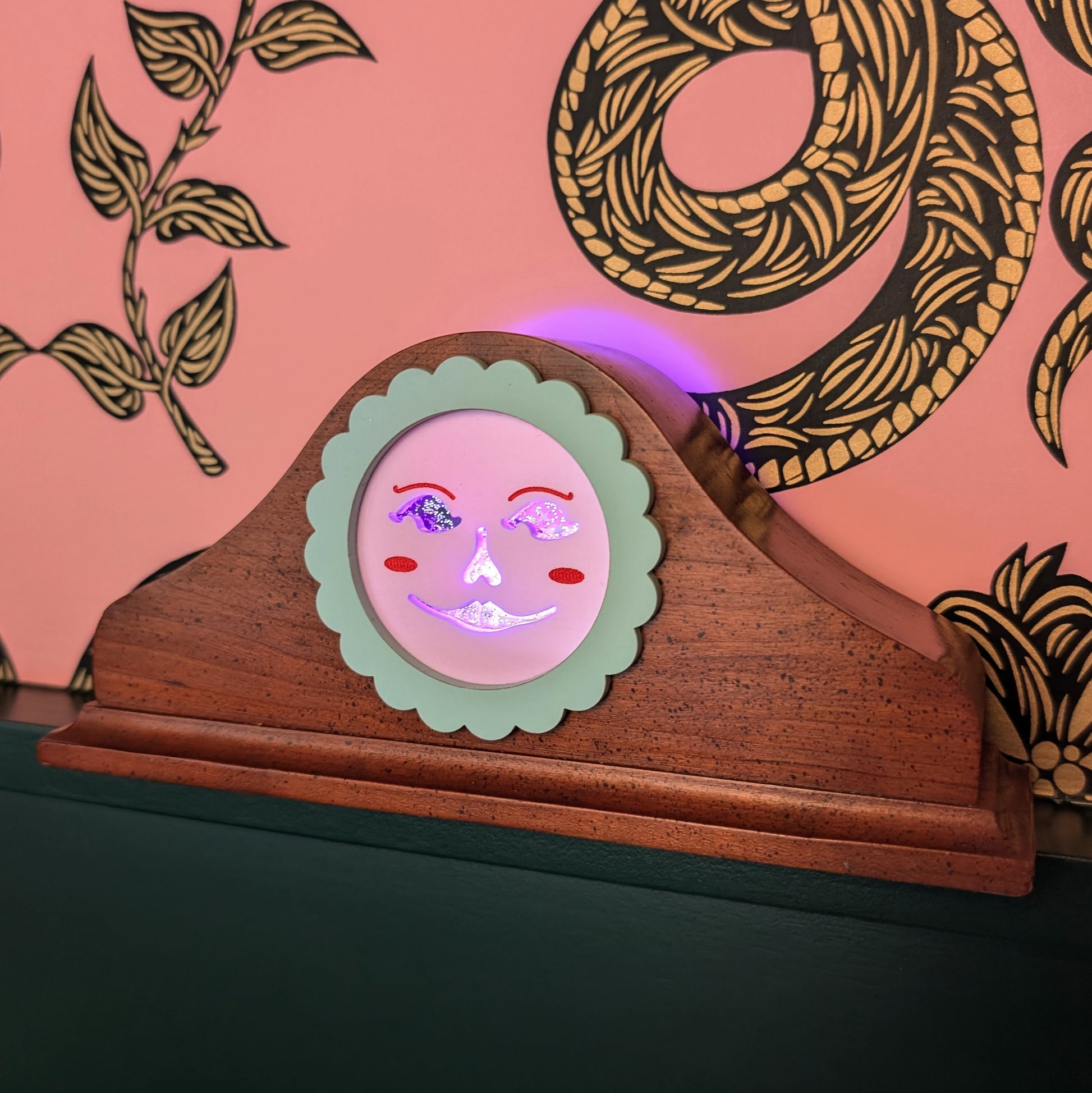 A vintage wood clock frame repurposed with a pink jack-o-lantern face. The face is lit up.