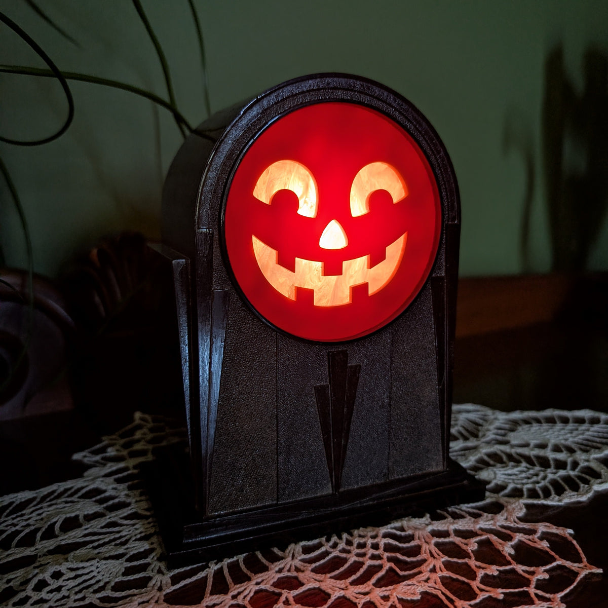 A vintage art deco themed plastic clock frame that has a cheerful jack-o-lantern face is sitting on a doily against a green wall. The scene is dark with the face lit up from inside.