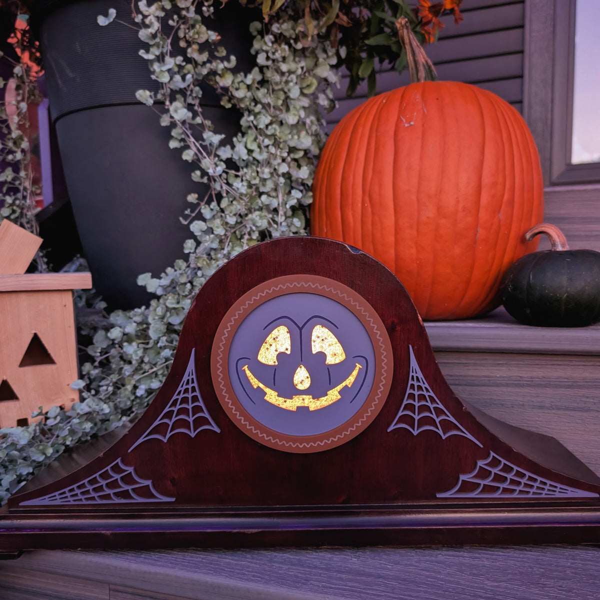 A vintage wood clock frame that has a grinning jack-o-lantern for the face and is sitting on a step against a plants and pumpkins The scene is dark with the face lit up from inside.