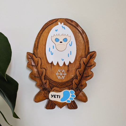 A leaf and acorn wooden plaque with an acrylic yeti face mounted in the middle.