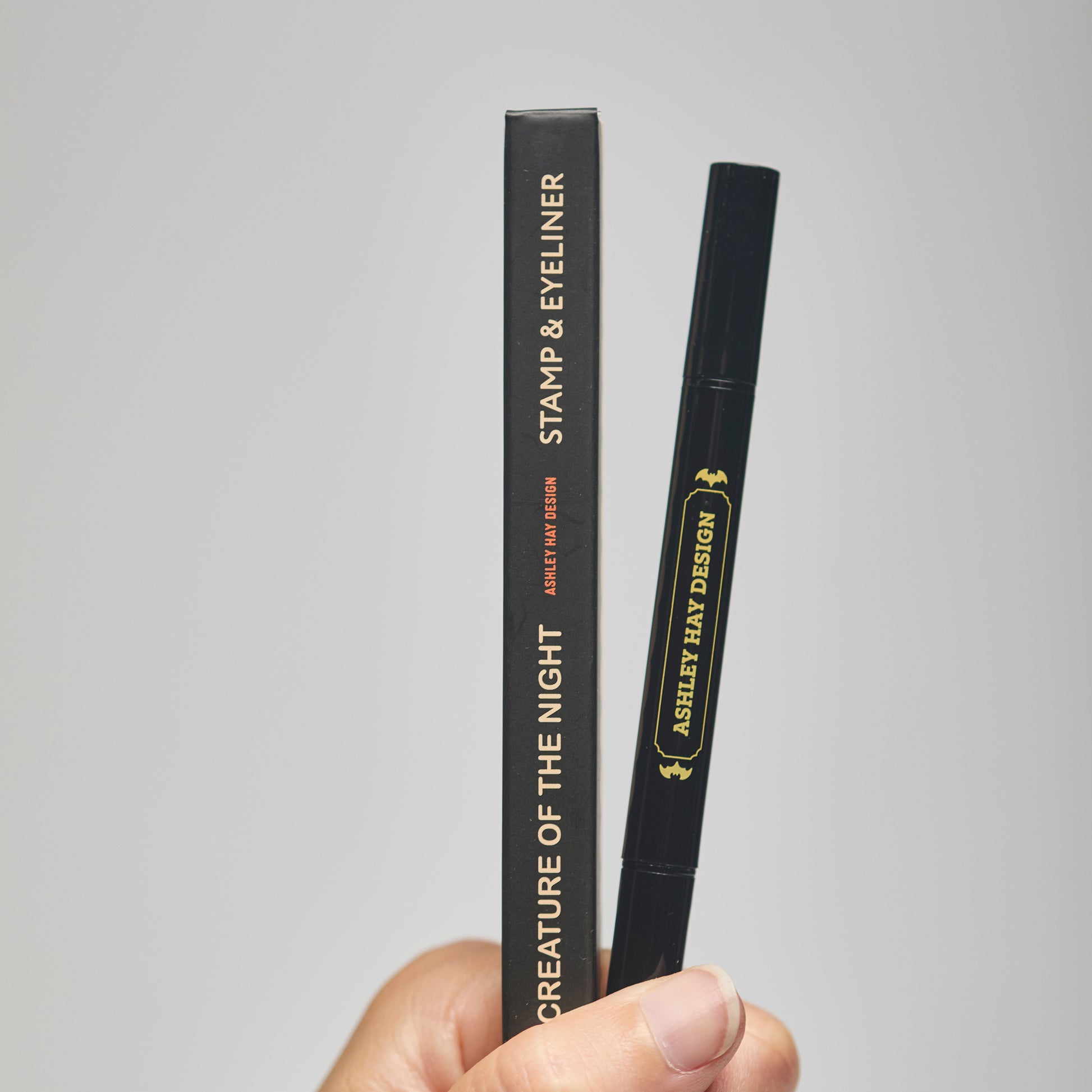 Hand holding a black makeup stamp and eyeliner package with visible branding against a neutral background