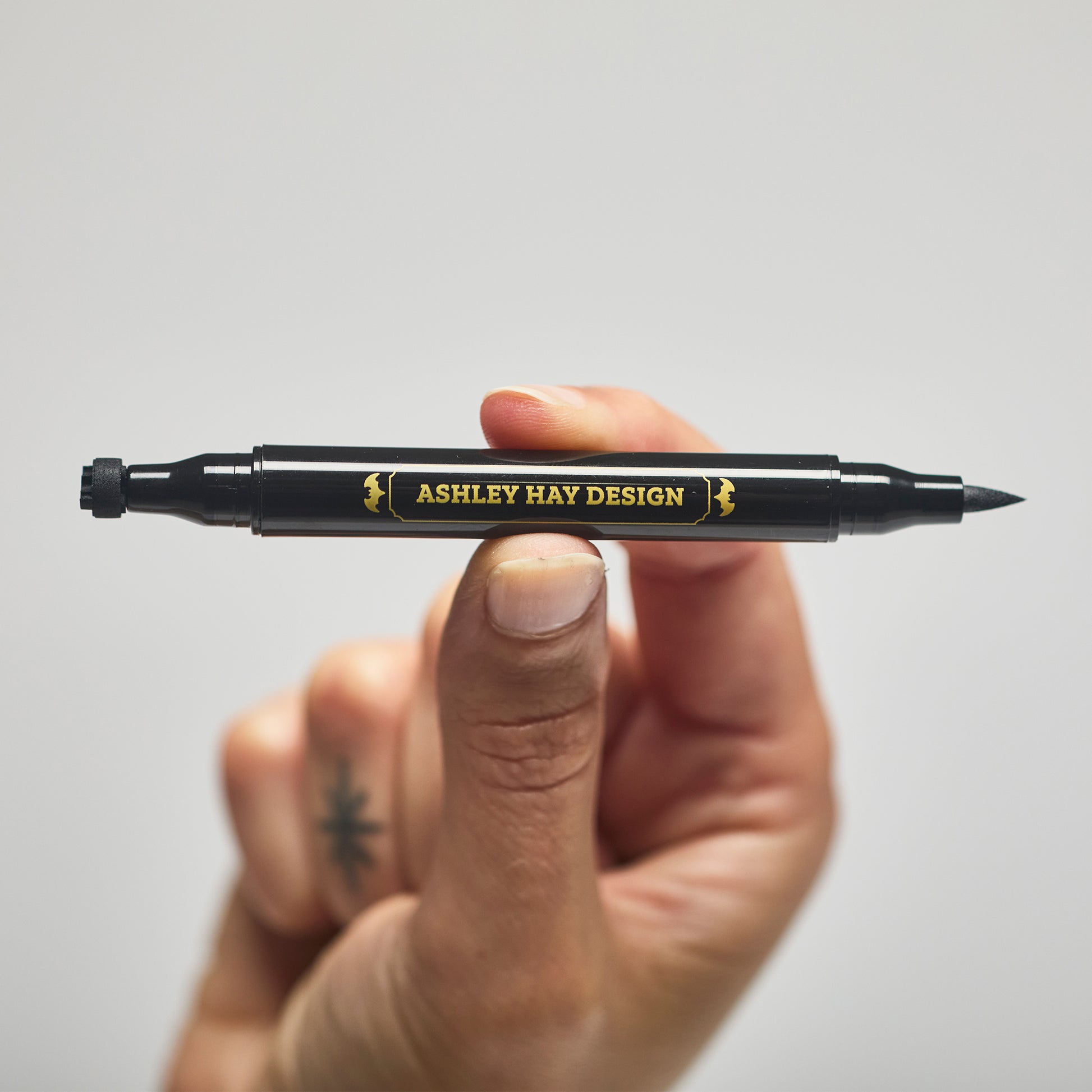 Hand holding a black makeup tube with a brush on one side and a stamp on the other side against a gray background.