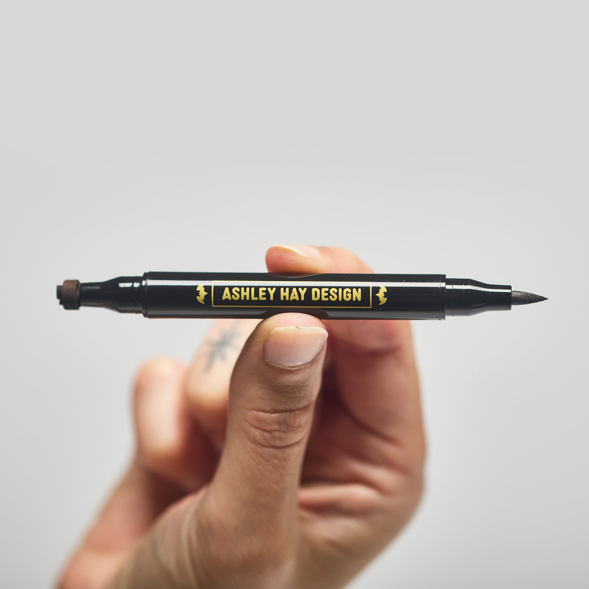 Hand holding a black makeup tube with a brush on one side and a stamp on the other side against a gray background.