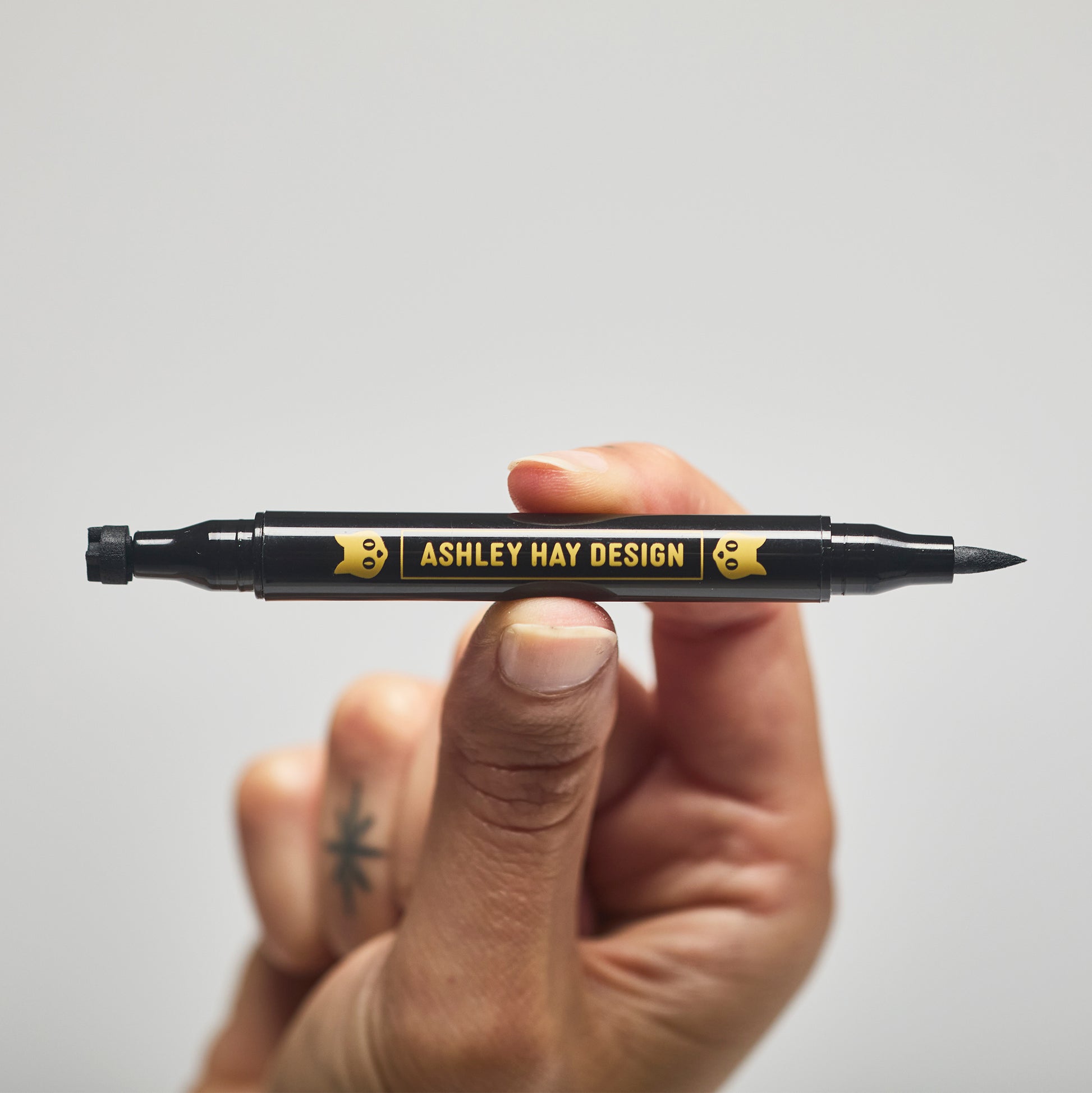 Hand holding a black makeup tube with a brush on one side and a stamp on the other side against a gray background.