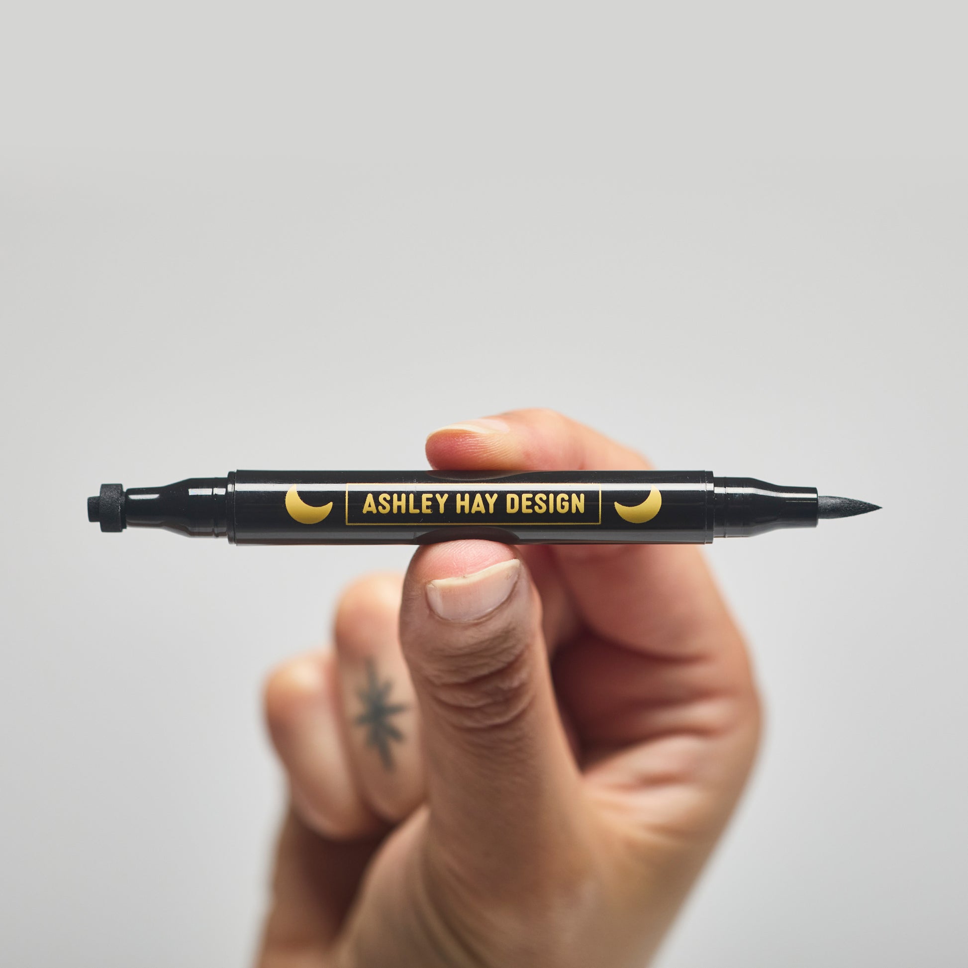 Hand holding a black makeup tube with a brush on one side and a stamp on the other side against a gray background.