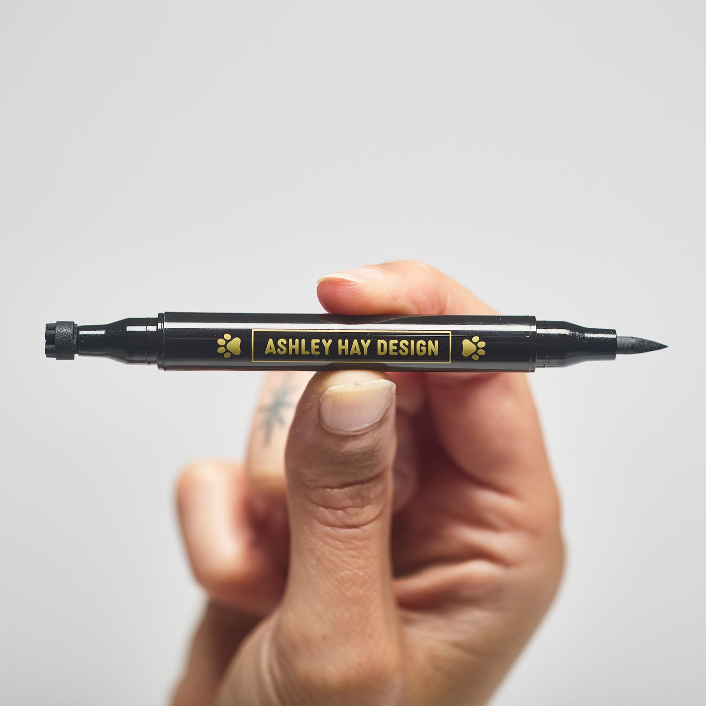 Hand holding a black makeup tube with a brush on one side and a stamp on the other side against a gray background.