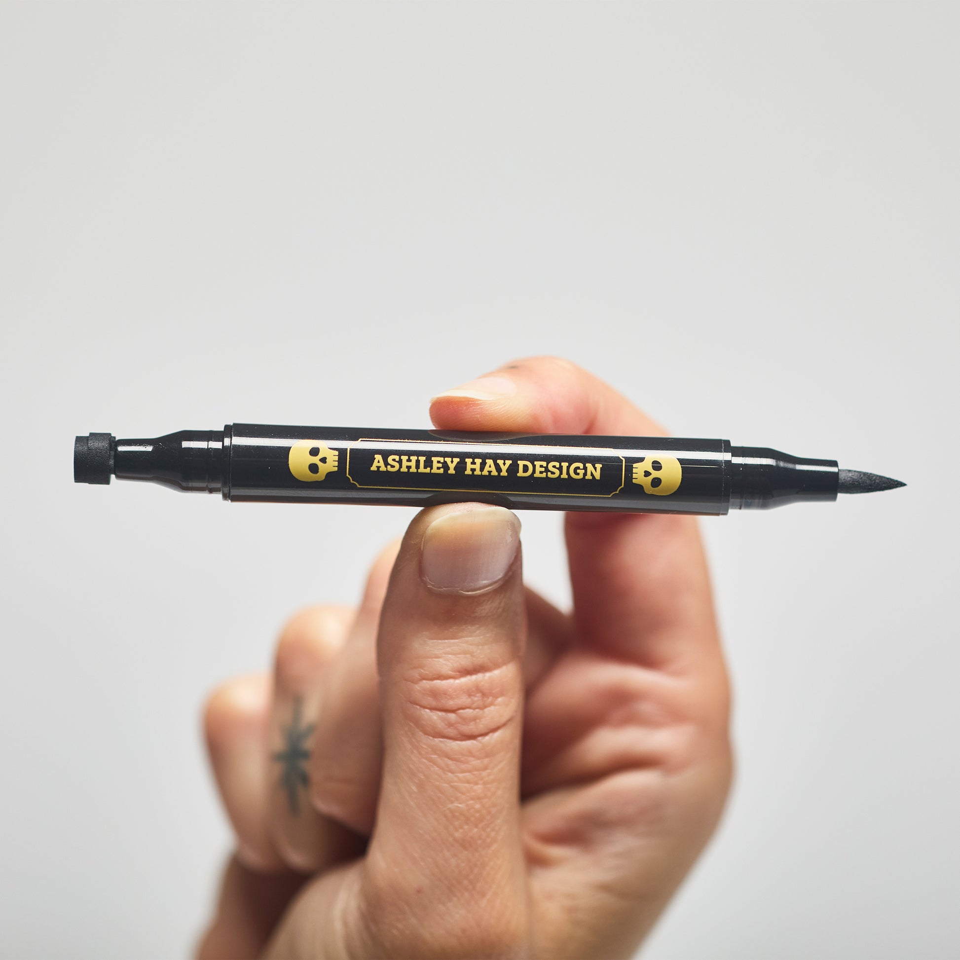 Hand holding a black makeup tube with a brush on one side and a stamp on the other side against a gray background.
