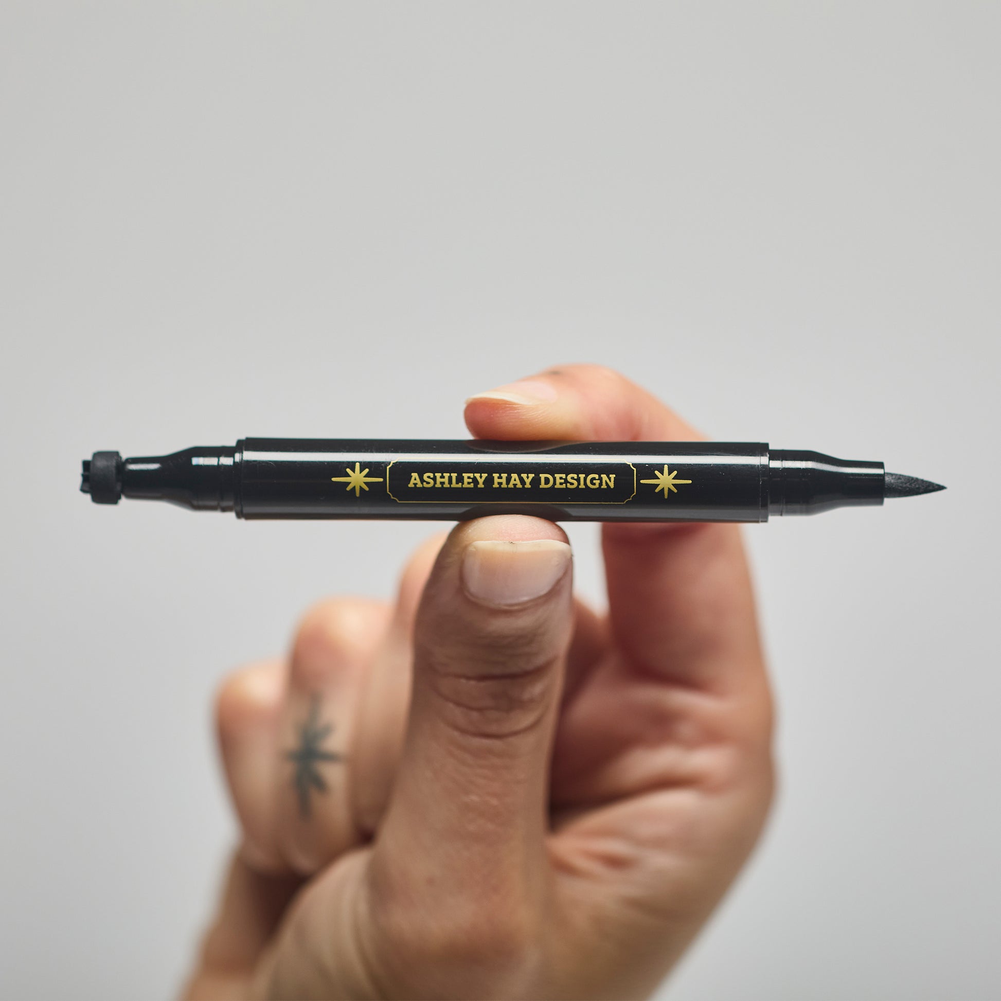 Hand holding a black makeup tube with a brush on one side and a stamp on the other side against a gray background.
