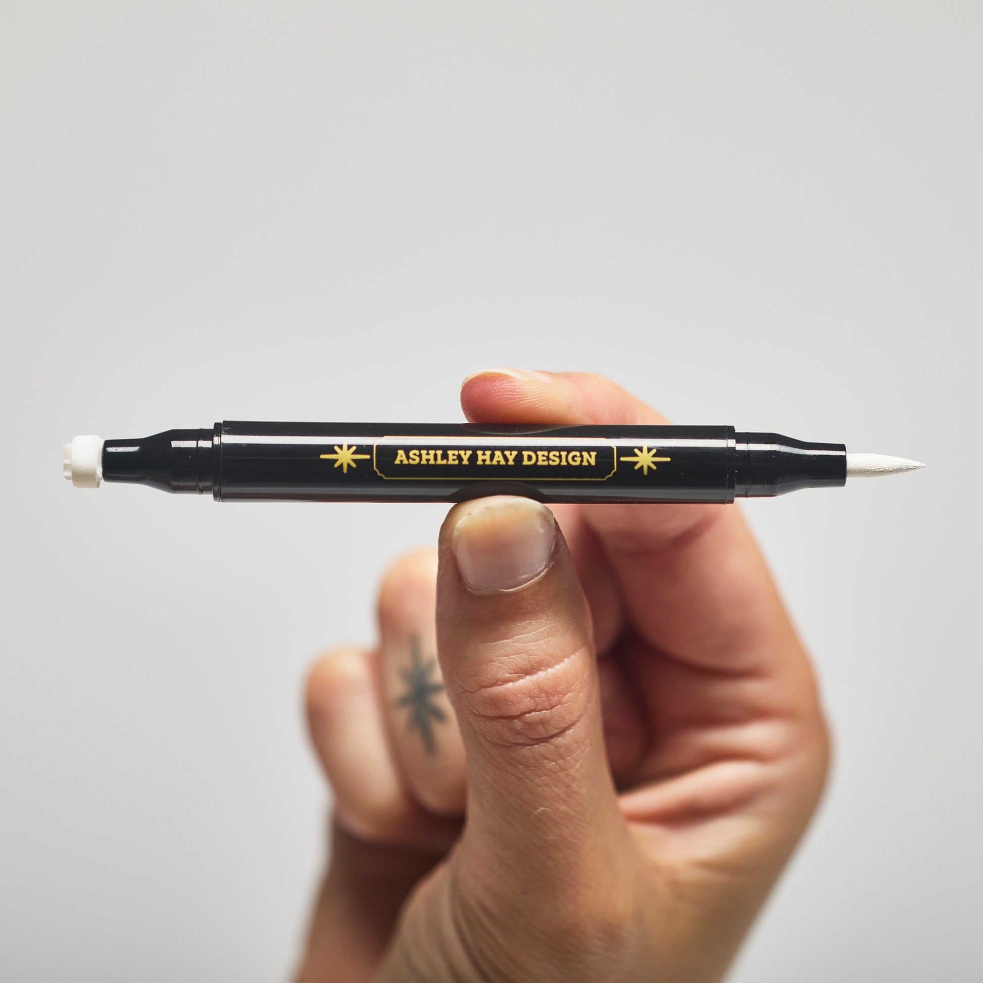 Hand holding a black makeup tube with a white ink brush on one side and a stamp on the other side against a gray background.