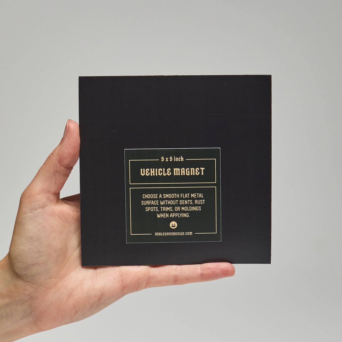 Hand holding a black square vehicle magnet packaging with text that reads '5 x 5 inch. Vehicle magnet. Choose a smooth flat metal surface without dents, rust, spots, trims, or moldings when applying.'
