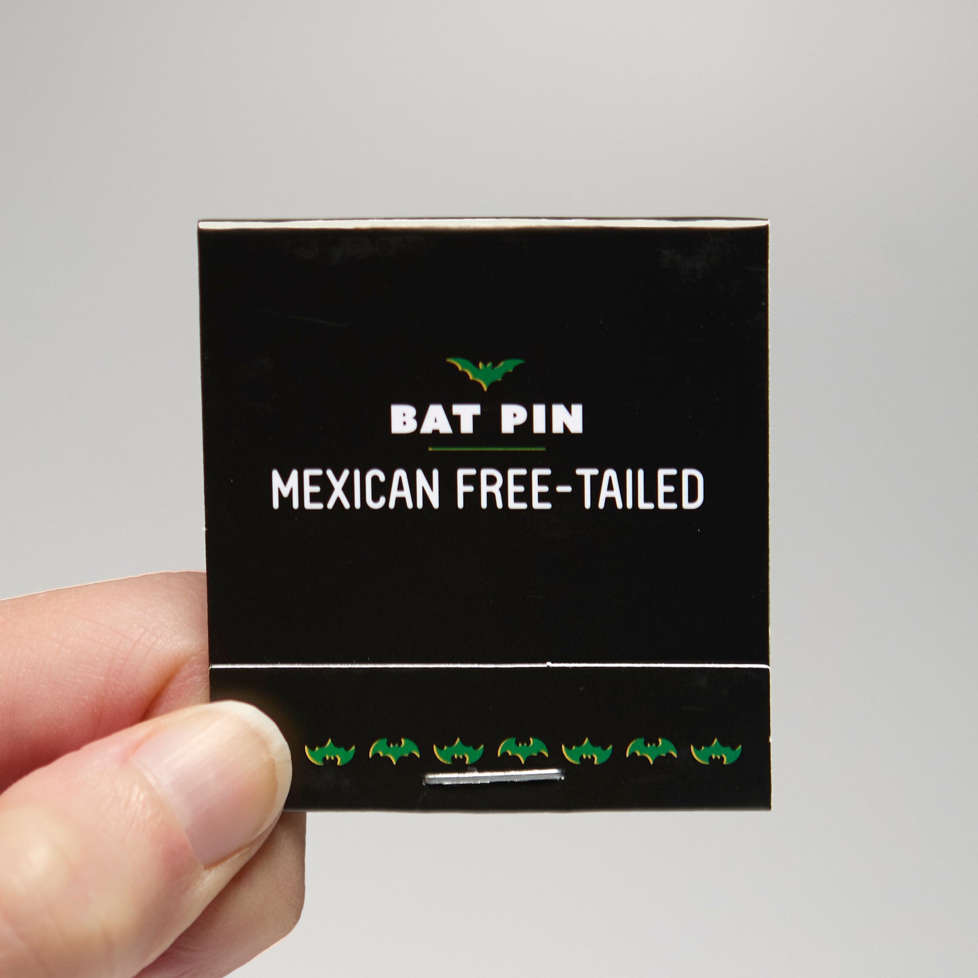 A black match book with 'Bat Pin Mexican free-tailed' text held by a hand against a neutral background