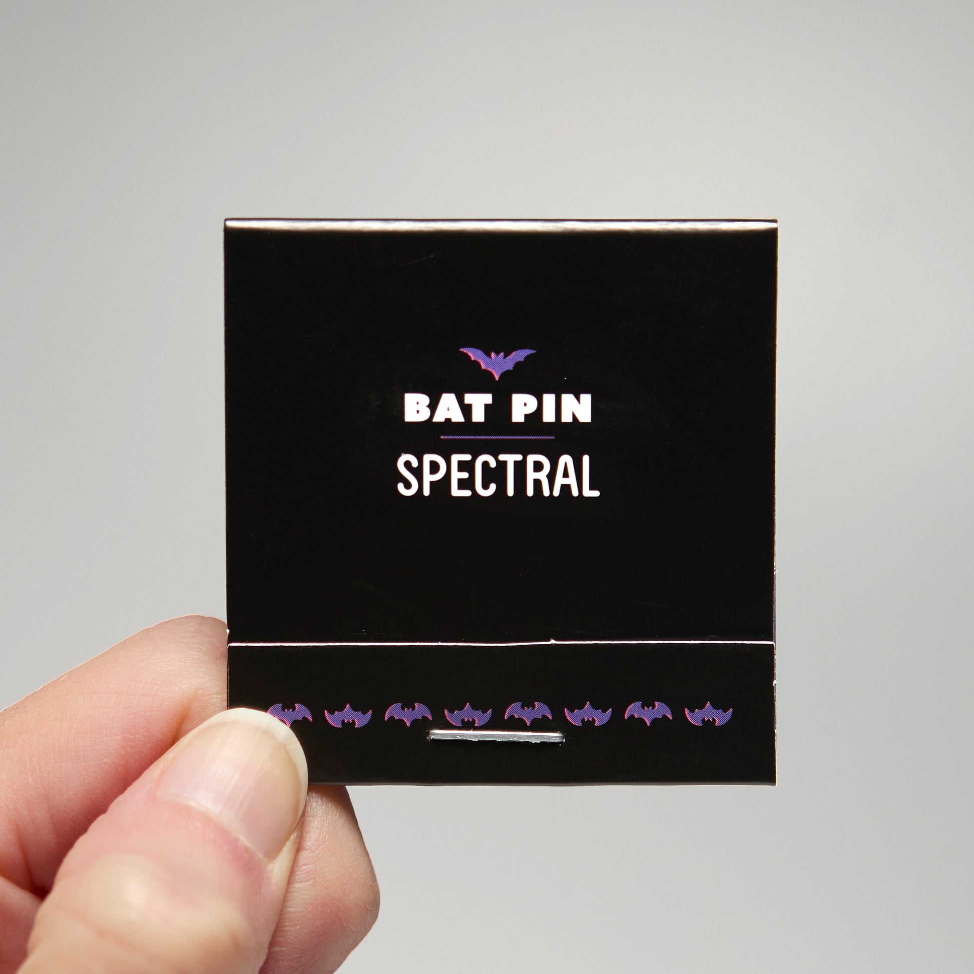 A black match book with 'Bat Pin Spectral' text held by a hand against a neutral background