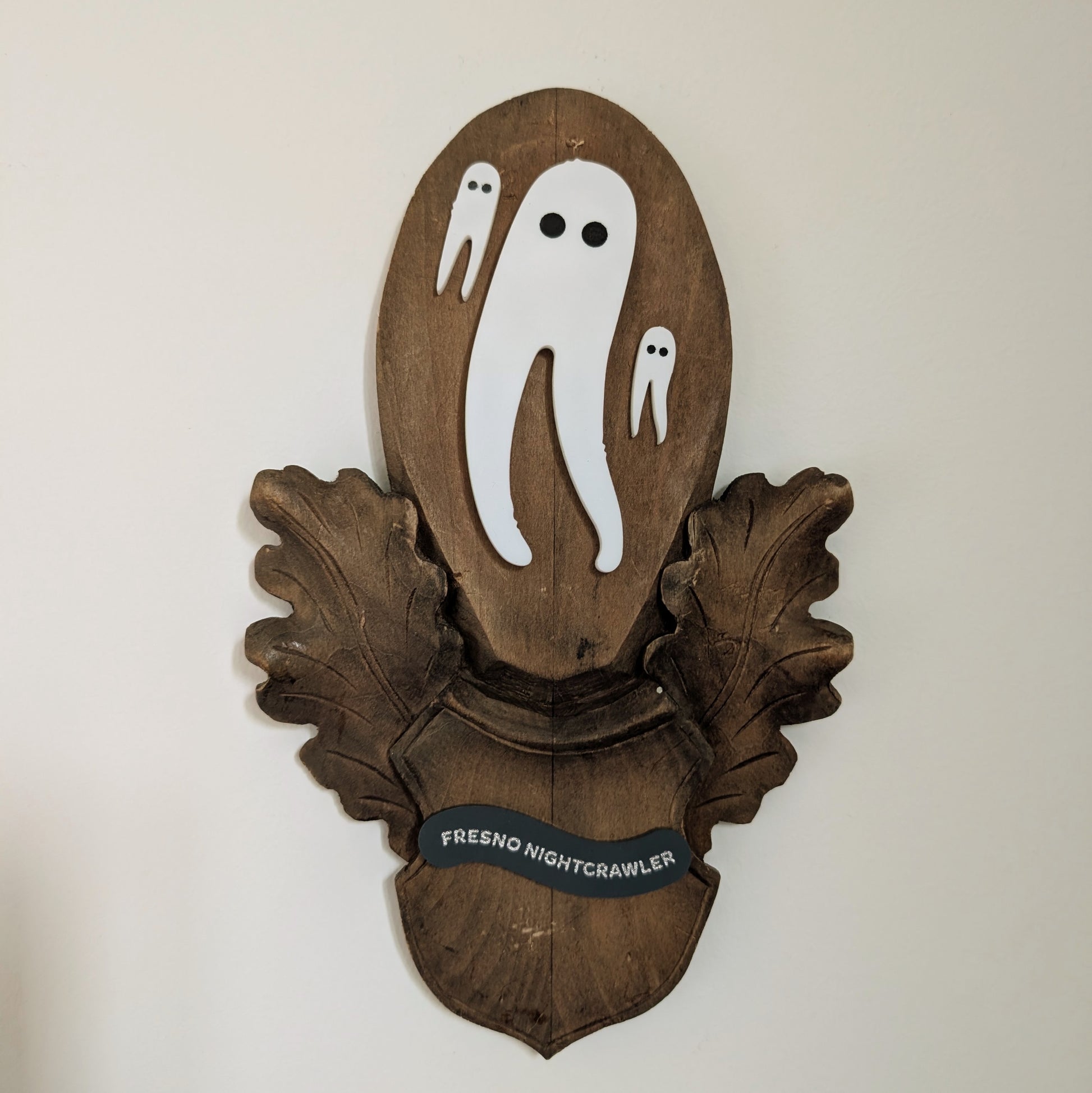 Three acrylic Fresno Nightcrawlers (white leggy cryptids are mounted on a vintage wooden plaque that hangs on a white wall.