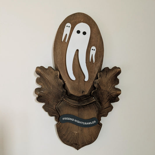Three acrylic Fresno Nightcrawlers (white leggy cryptids are mounted on a vintage wooden plaque that hangs on a white wall.